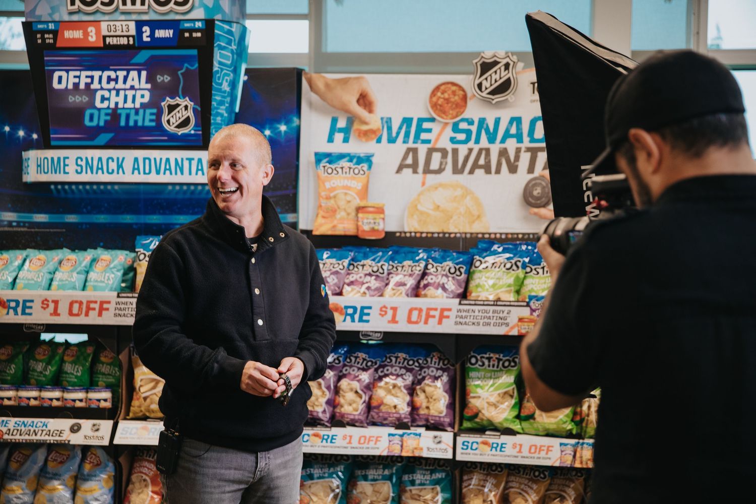 A man smiles during a commercial shoot in a convenience store snack aisle with NHL branding displays.