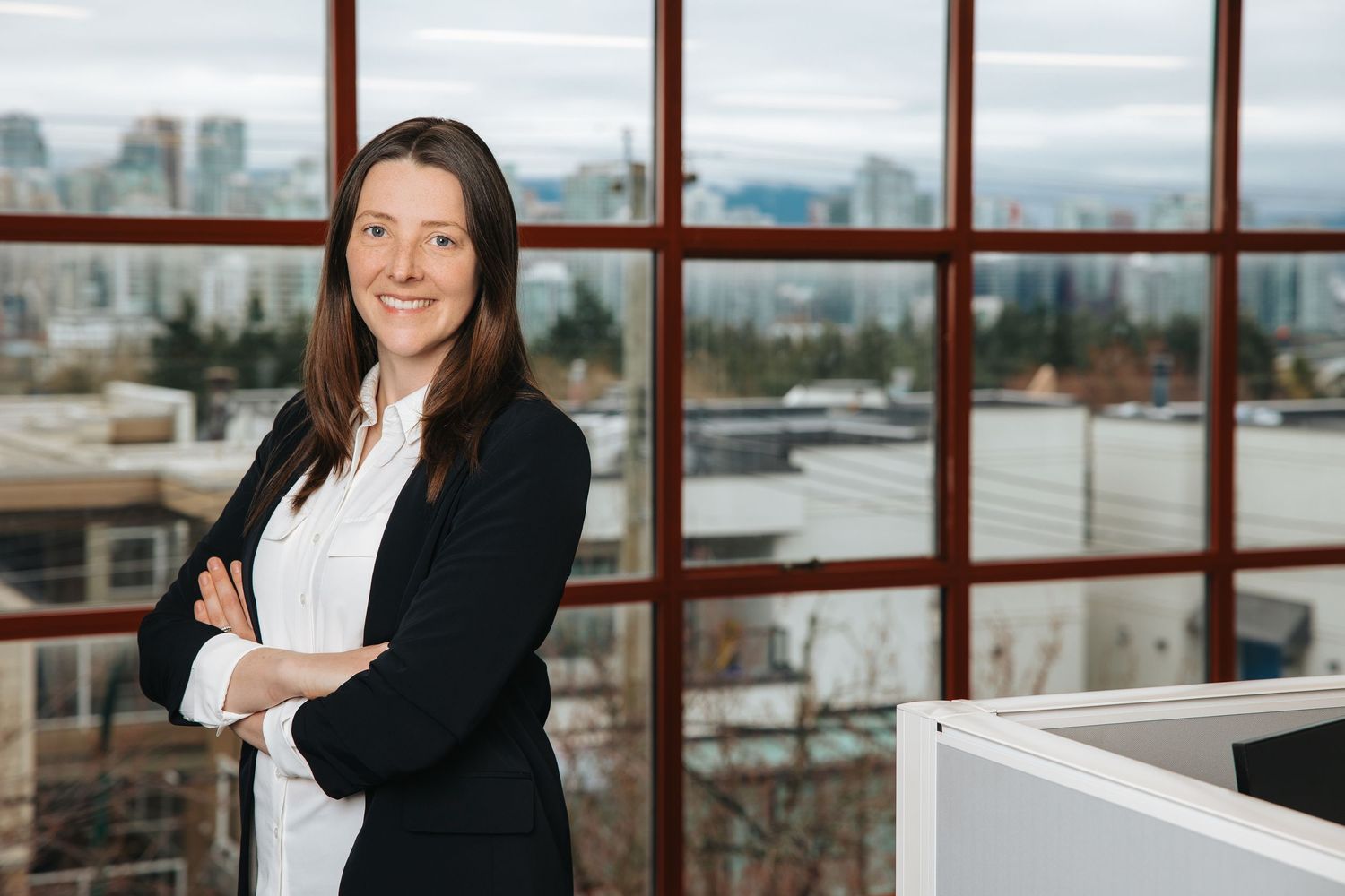 Professional in business attire stands by large office windows with cityscape views in background.