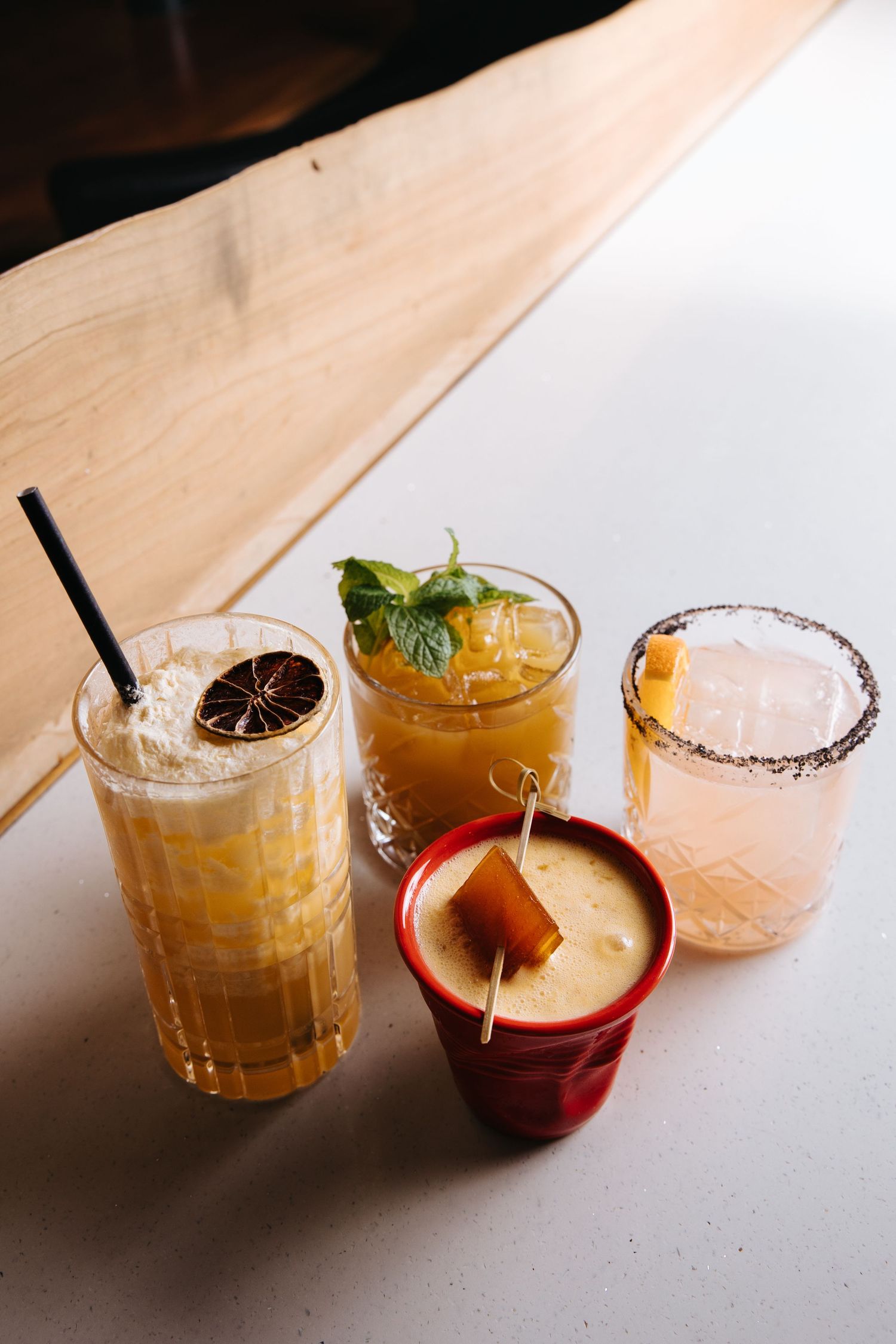 Four colorful cocktails with garnishes including mint, citrus, and fruit skewers arranged on a white surface.