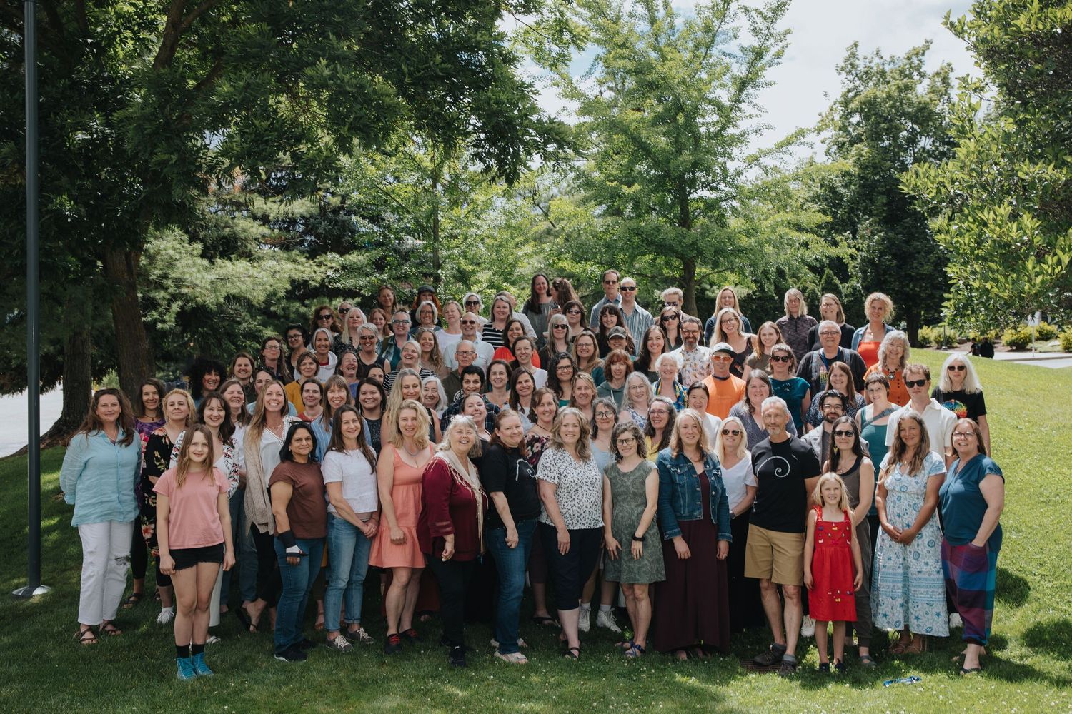 Large group of people gathered outdoors for a formal photo on a sunny day with trees in the background.