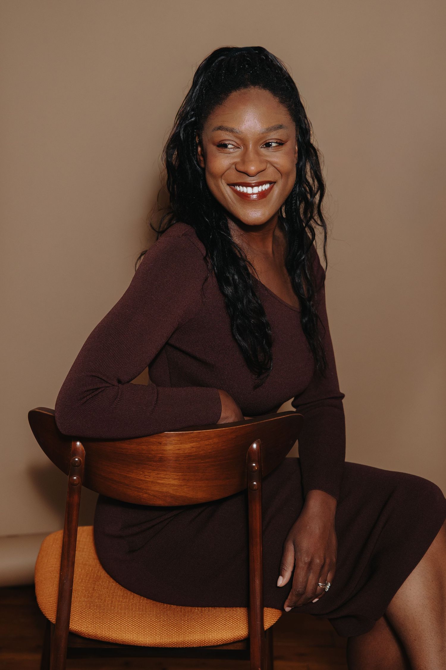 Portrait series in burgundy outfit seated on wooden chair against neutral backdrop.