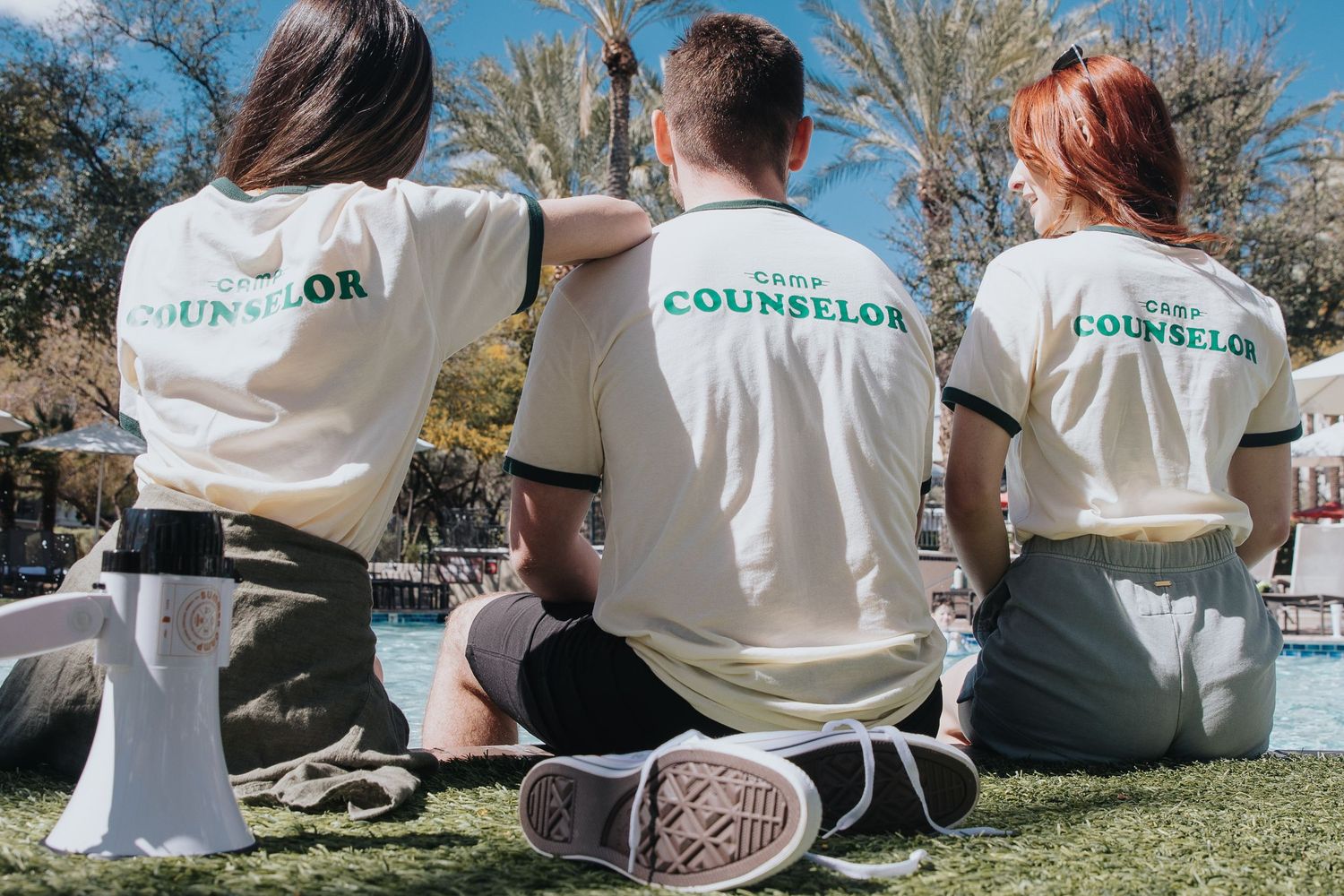 Row of camp counselors sitting together wearing matching white shirts by the pool.