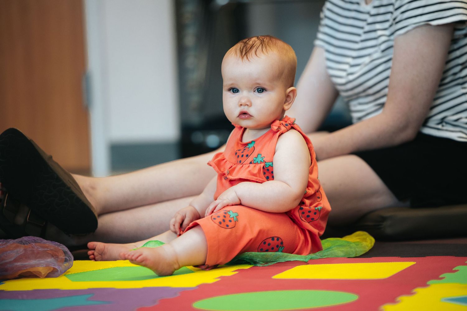 Baby in orange outfit sitting on colorful play mat during indoor activity.