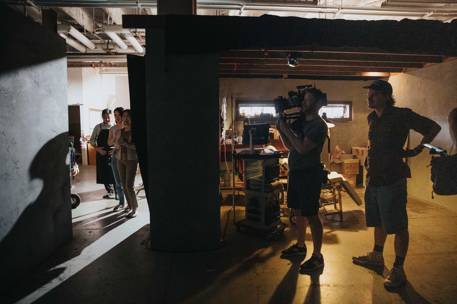 Behind-the-scenes film crew shooting in a dimly lit industrial studio space with camera operator and director.