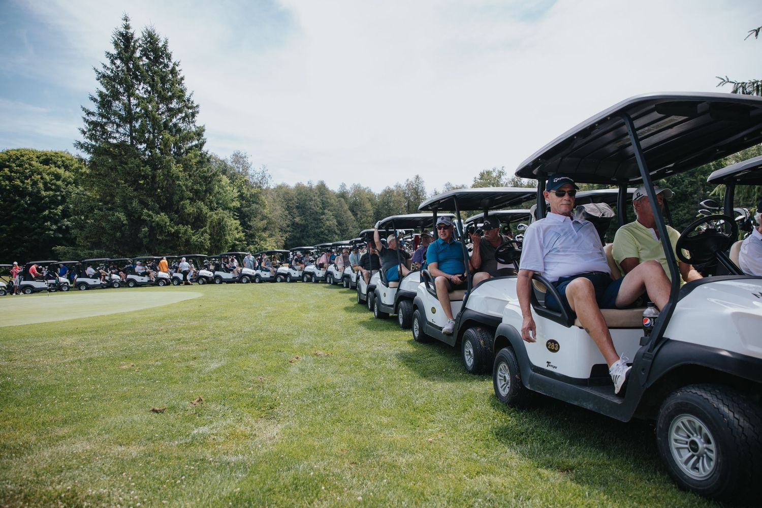 Rows of golf carts lined up on a green course with players seated, surrounded by tall trees on a sunny day.