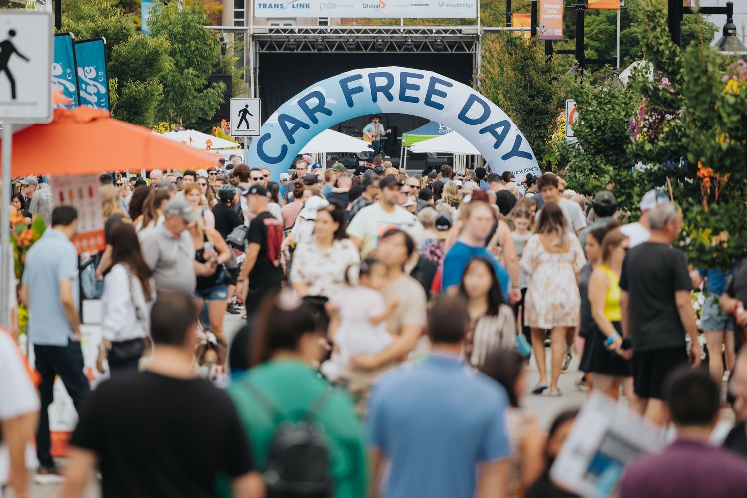 Large crowd enjoying a summer street festival with banners and trees.