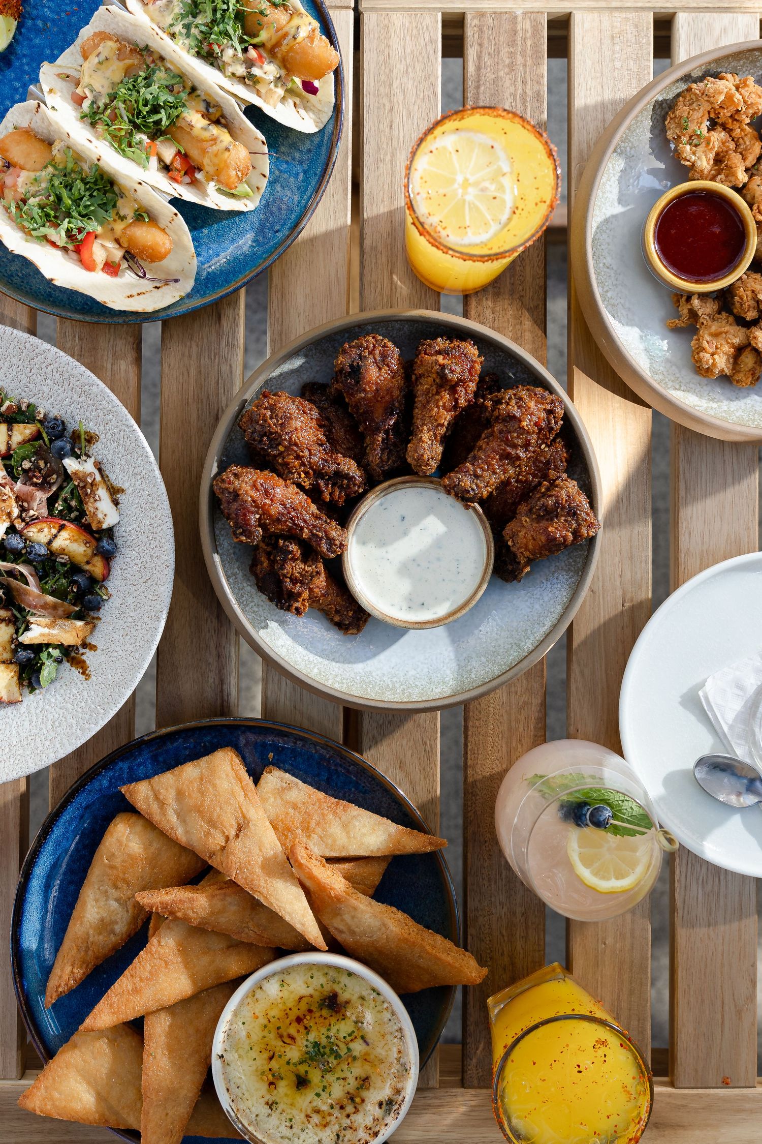 Overhead view of a restaurant spread featuring tacos, fried chicken, samosas, hummus, and lemon cocktails on wooden table.