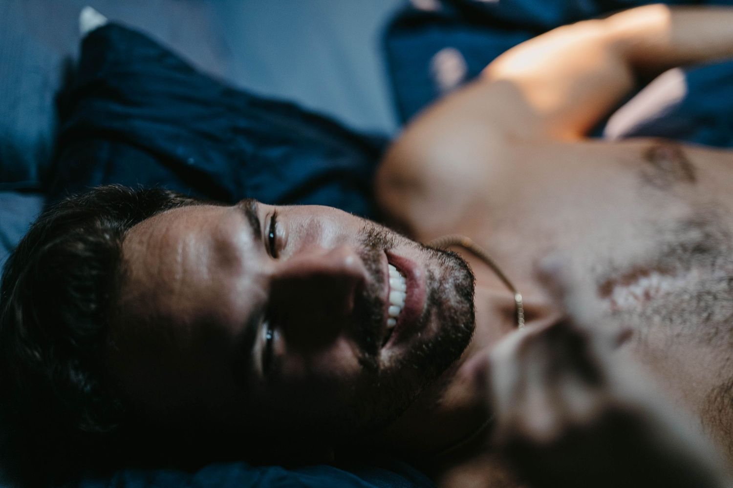 Smiling bearded man lying on blue bedding with eyes closed in relaxed peaceful pose.