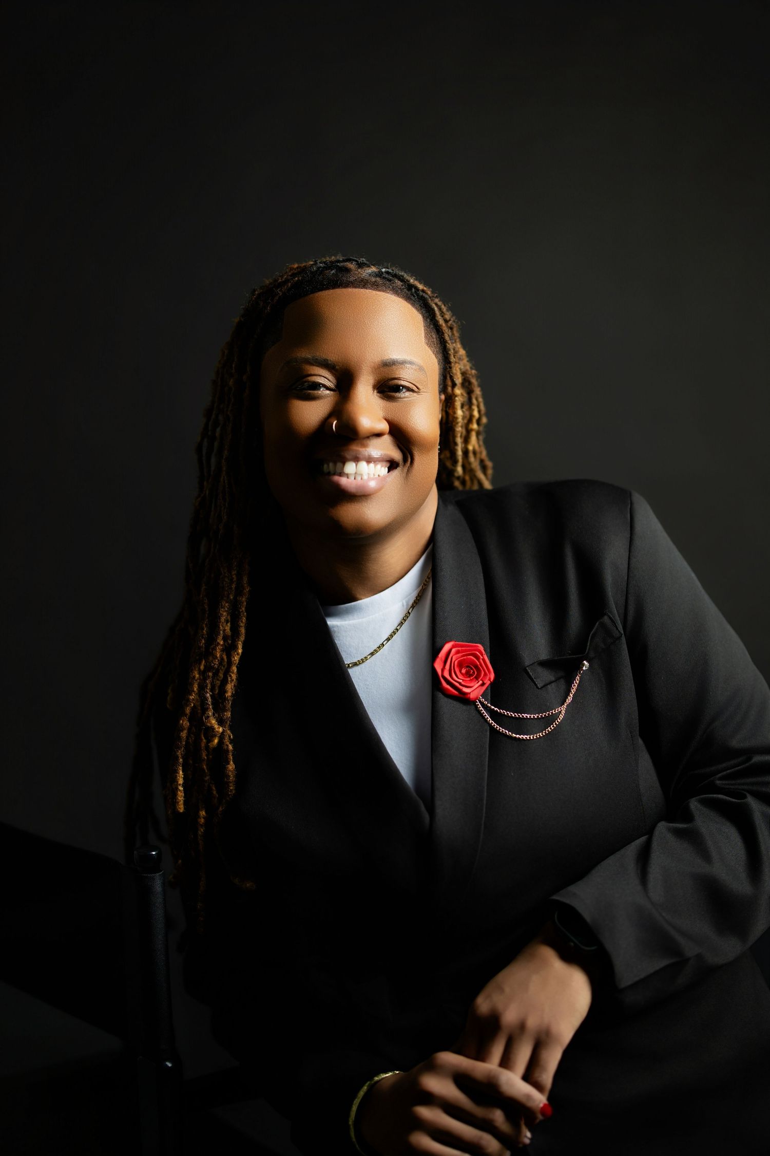 Professional studio portrait against dark background featuring warm smile, dreadlocks, black blazer and red lapel flower.