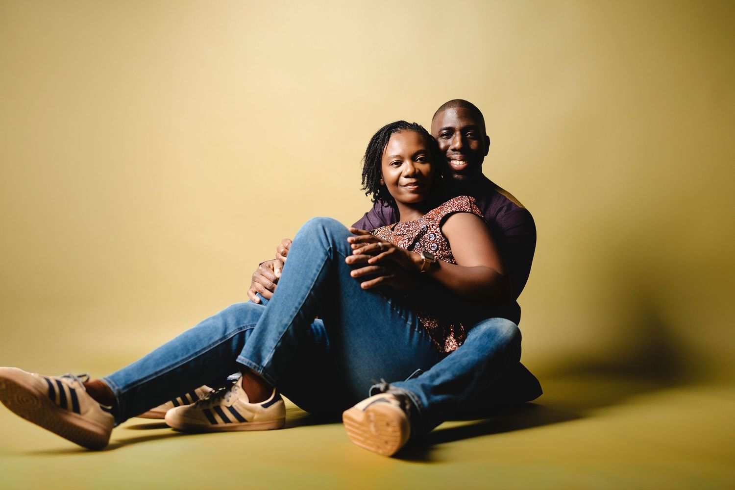 A couple sits together on a yellow studio floor, smiling warmly, with the woman leaning back into the man's embrace.