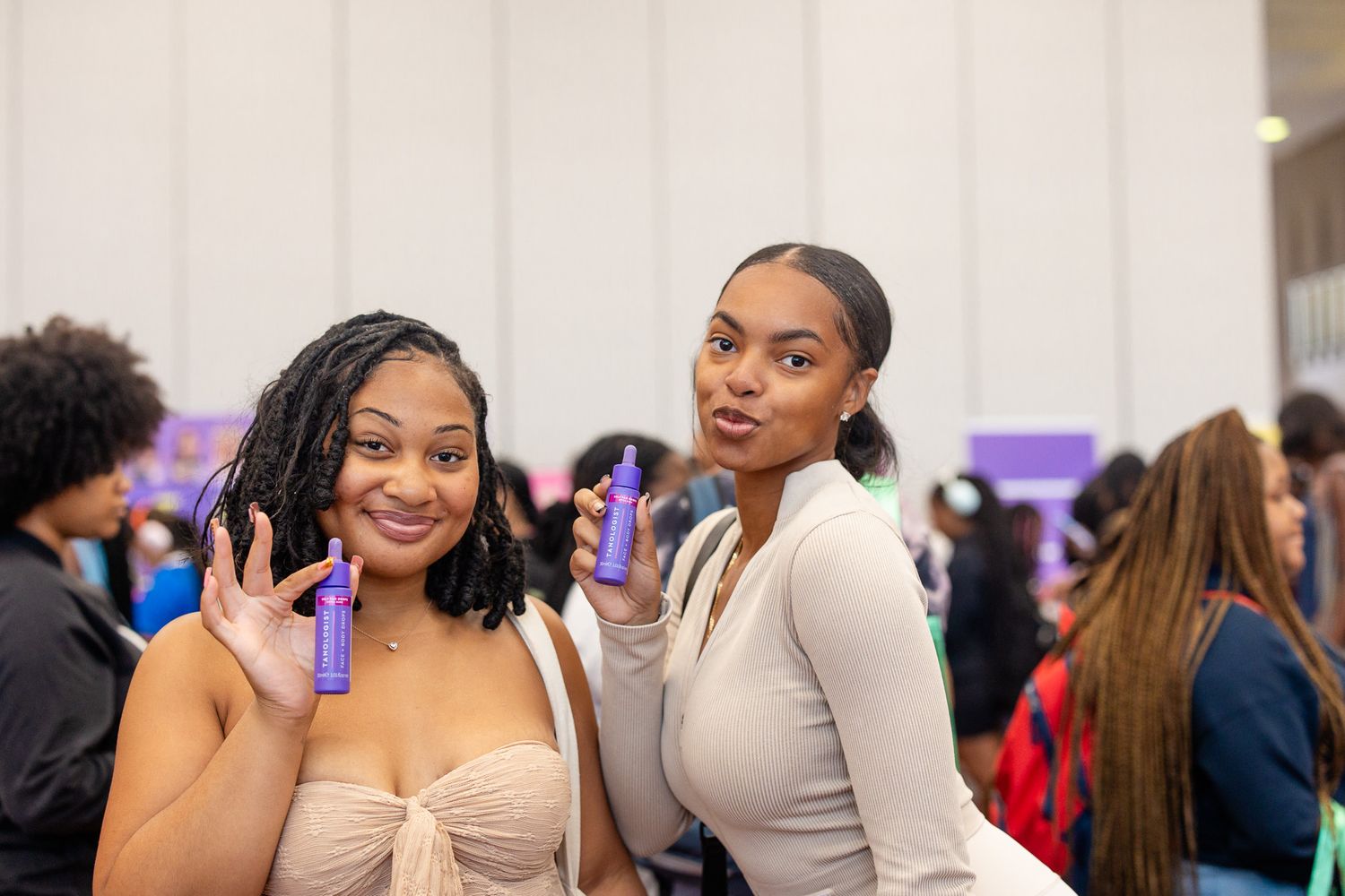 Two young women smiling and holding purple hair care products at a beauty expo event.