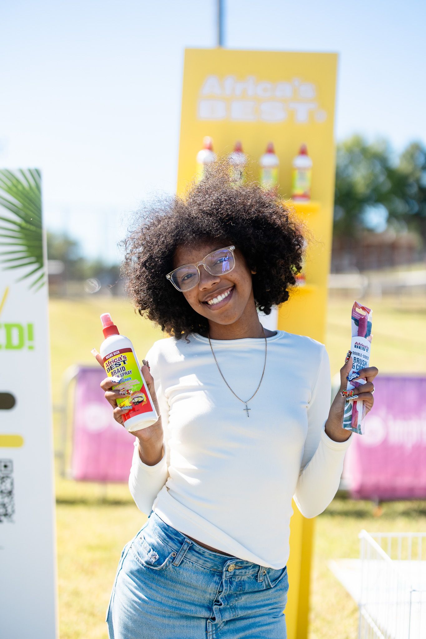Smiling young woman with curly hair holding Africa's Best hair products at an outdoor event.