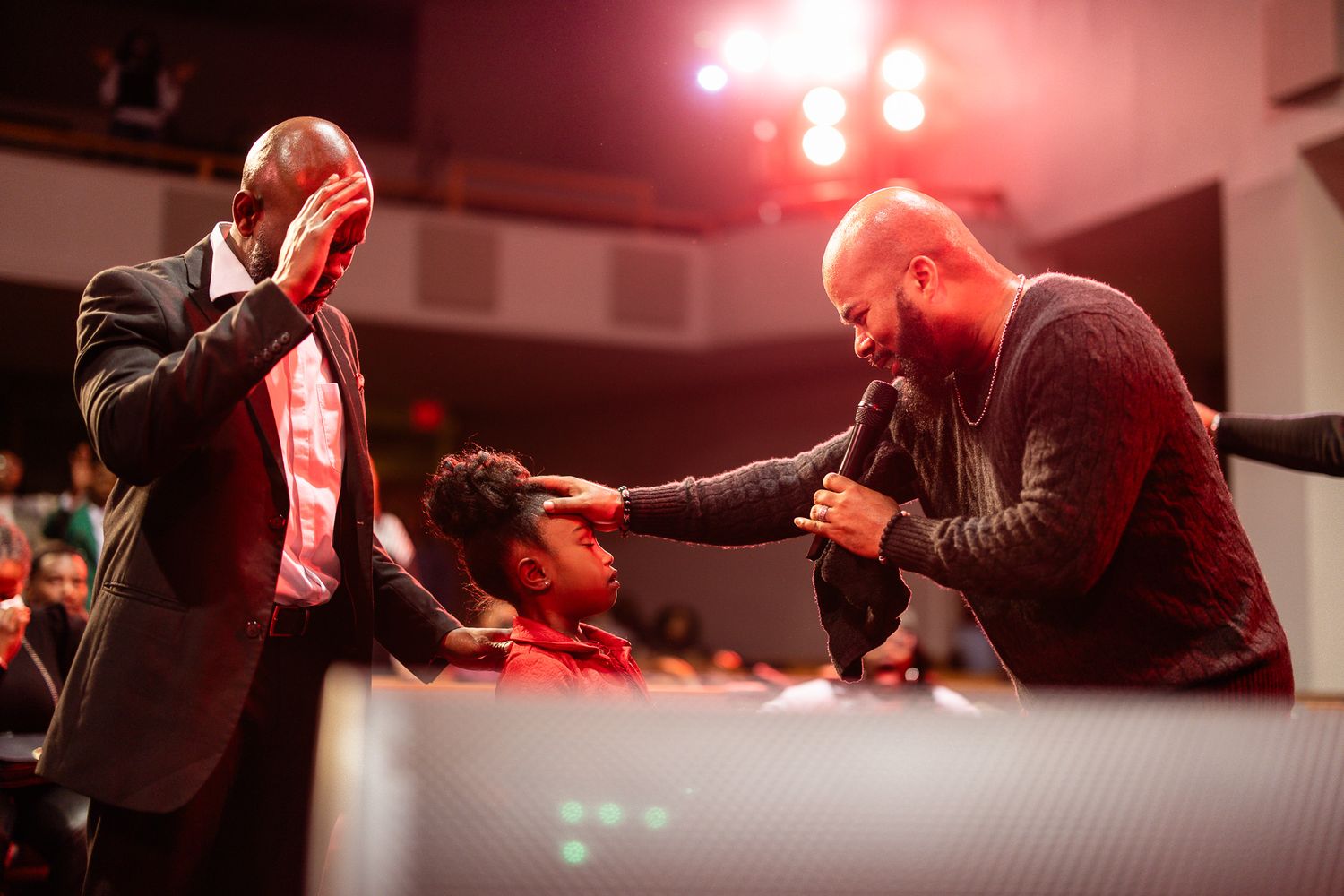 Two men pray over a child during a church service, with a congregation visible in the background.