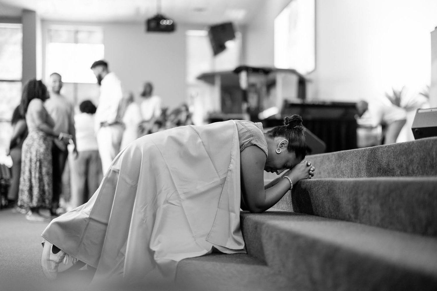 Black and white photo of a bride kneeling and praying in a church pew on her wedding day.