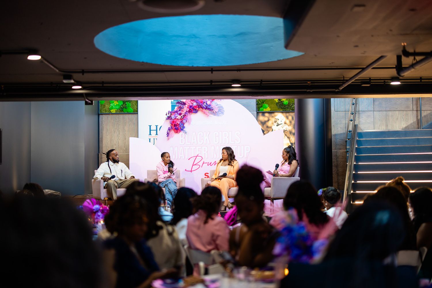 Panel discussion at an elegant event with speakers on stage before a floral backdrop, audience seated at decorated tables.