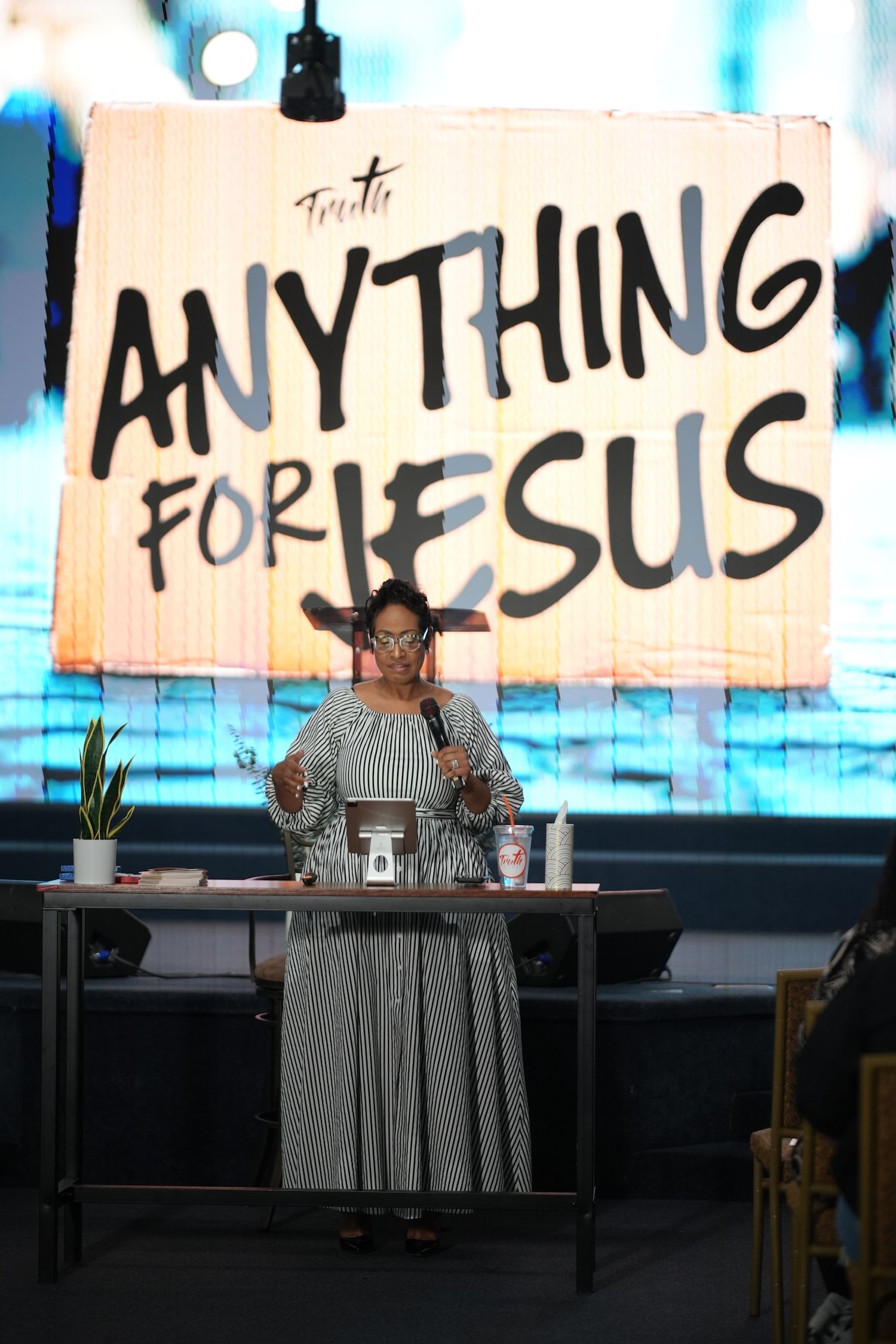 Woman speaking at a Christian event on stage with an Anything for Jesus banner displayed behind her.