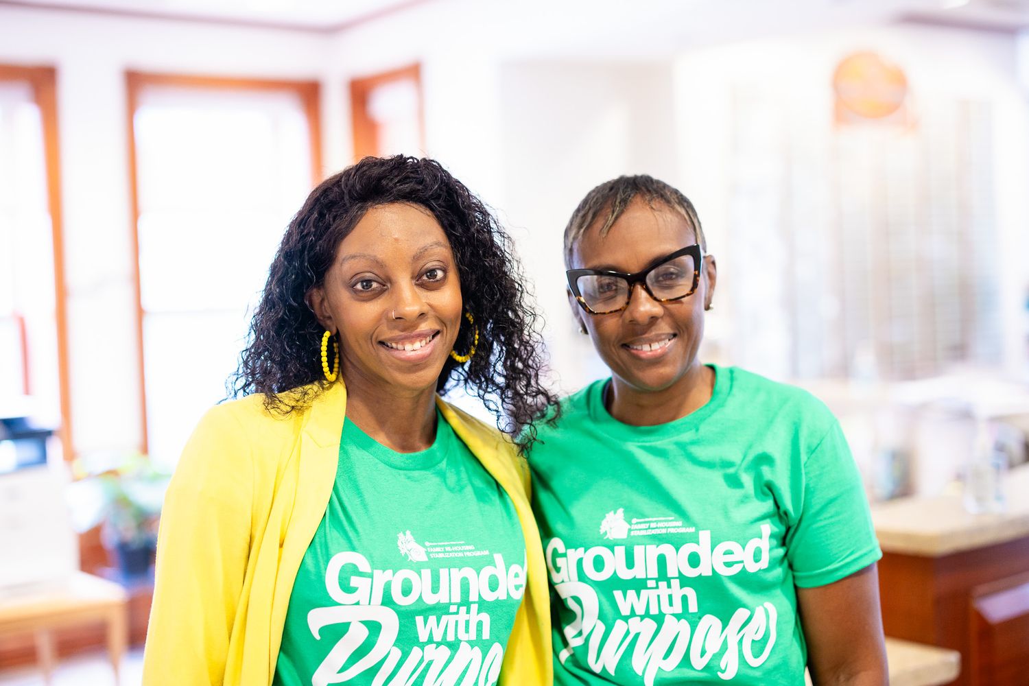 Two women smiling in green Grounded with Purpose t-shirts, posing together indoors.