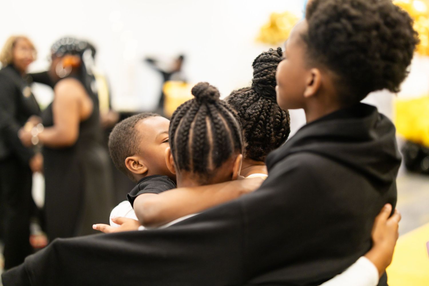 Three Black family members in black attire share a warm embrace at a gathering, with others visible in the background.