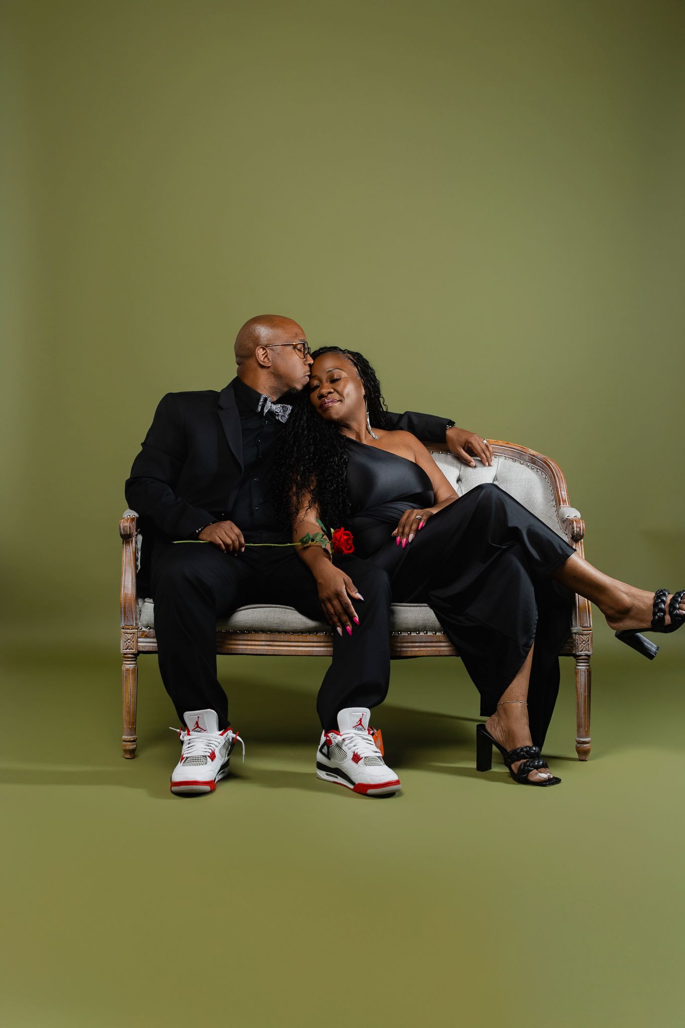 A couple sits intimately on an antique settee against a green backdrop, with him wearing sneakers and her in heels.
