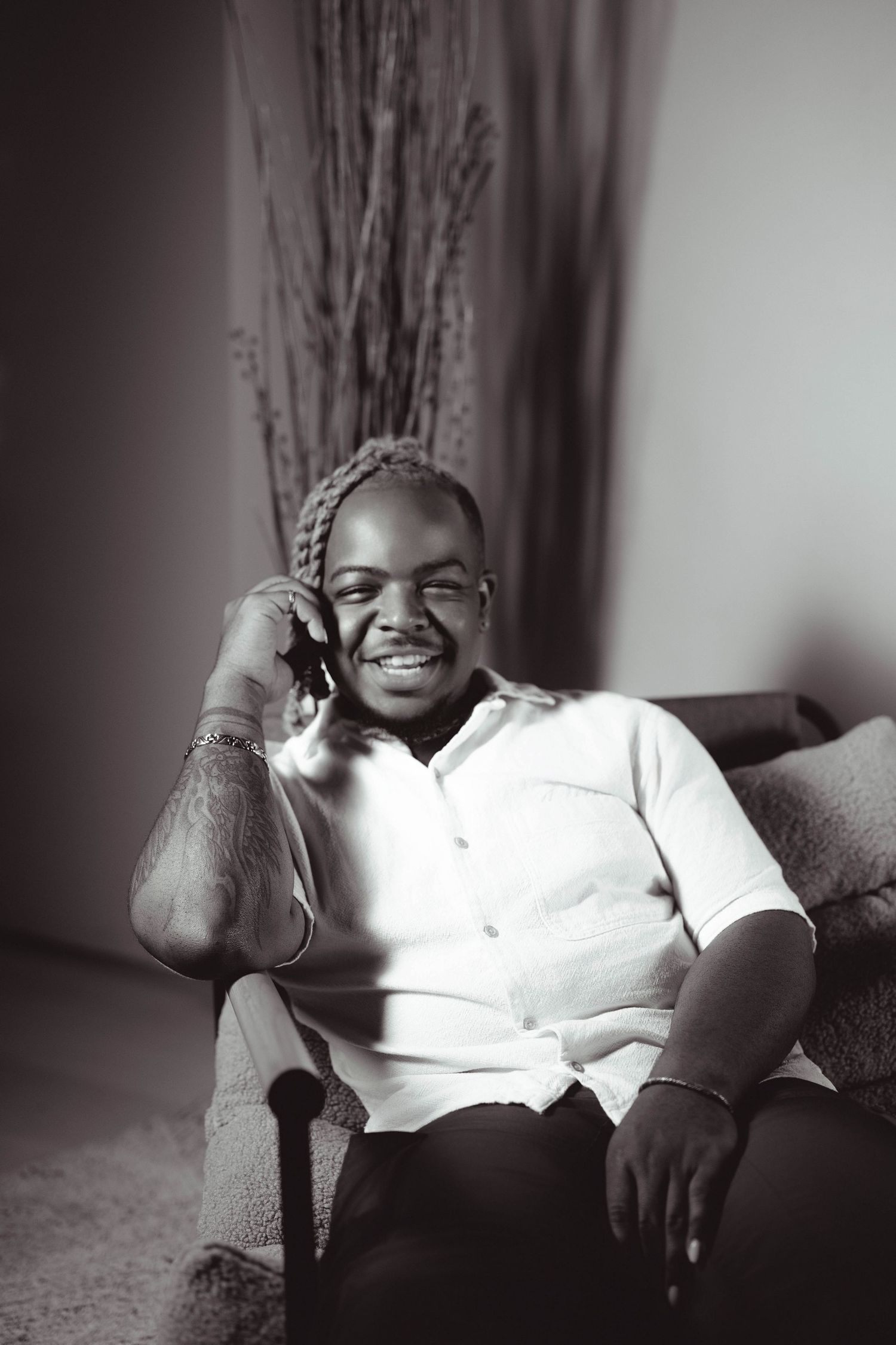 Black and white portrait of a smiling man talking on phone, seated in armchair wearing white shirt.