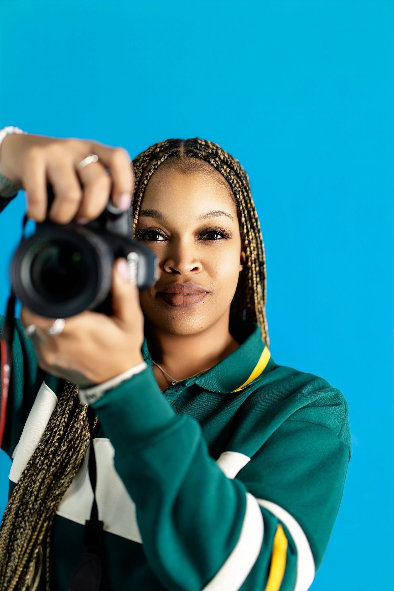 Young Black woman with braided hair holds a DSLR camera, wearing a green striped jacket against a blue background.