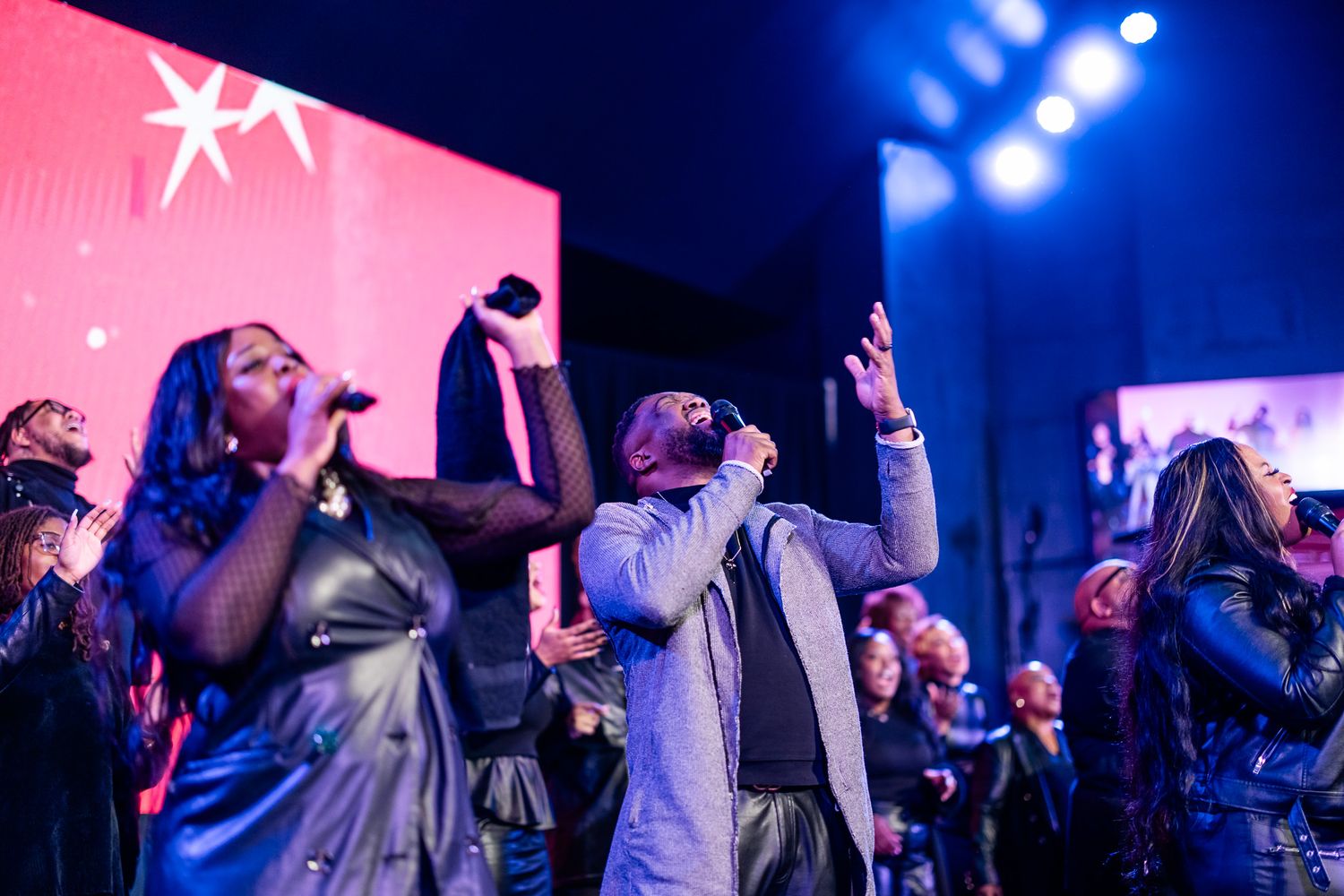 Gospel choir performing on stage with dramatic lighting, singers raising hands in worship during a live concert event.