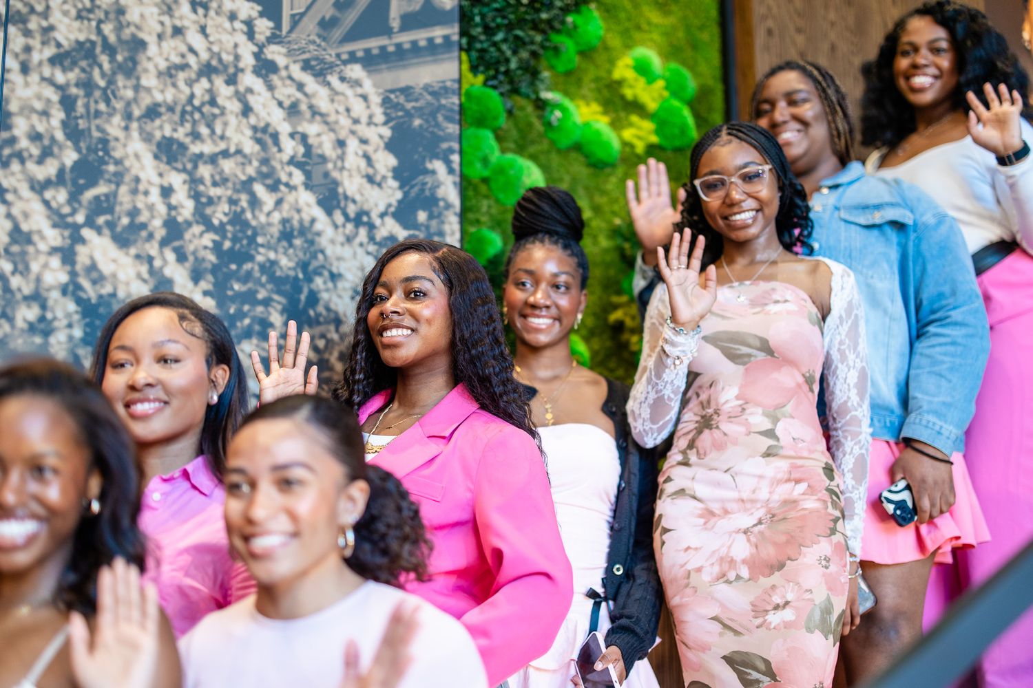 Group of young Black women in pink outfits smiling and waving at an outdoor celebration event.