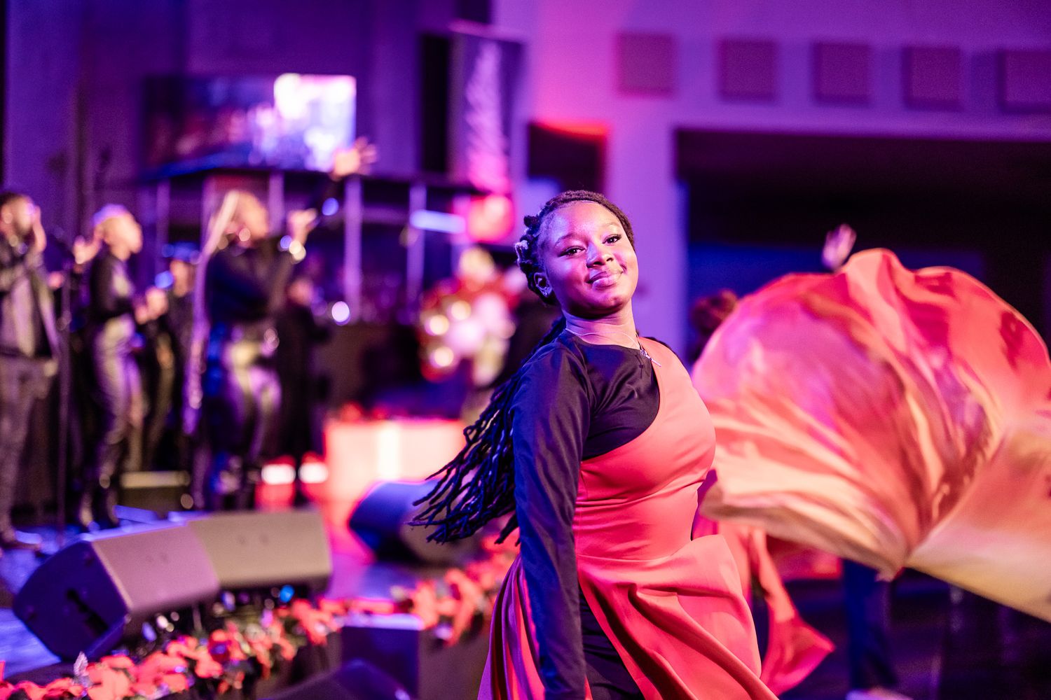 A woman in a red dress gracefully dances on stage with flowing fabric, performers visible in the background under purple lights.