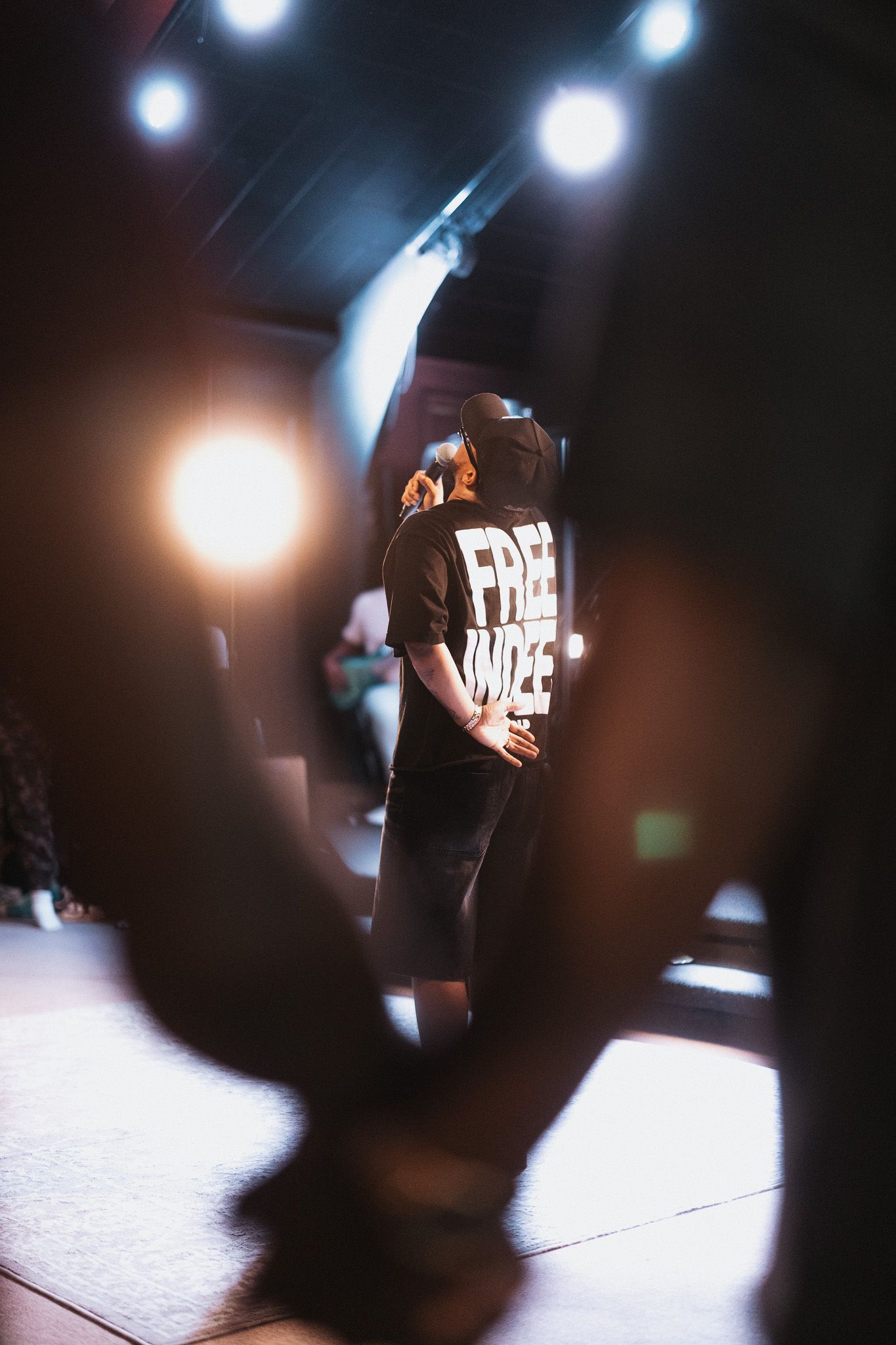 Person on stage wearing a black 'Free Wize' t-shirt under dramatic concert lighting.