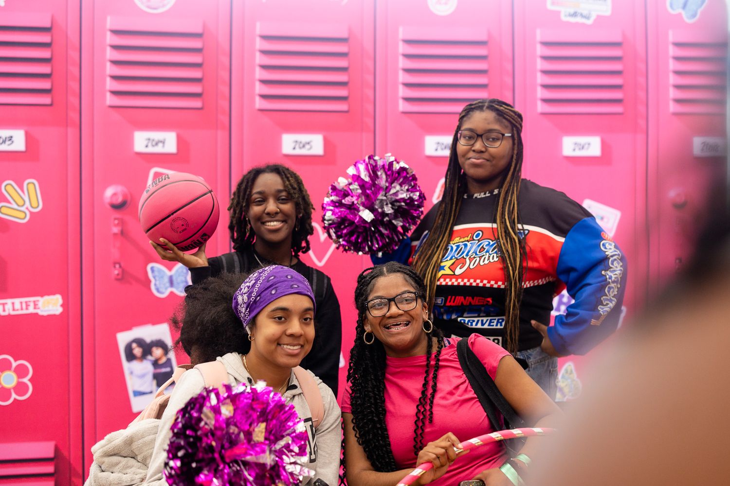 Four young Black girls pose cheerfully by pink lockers, holding pom-poms and a basketball in a school hallway.