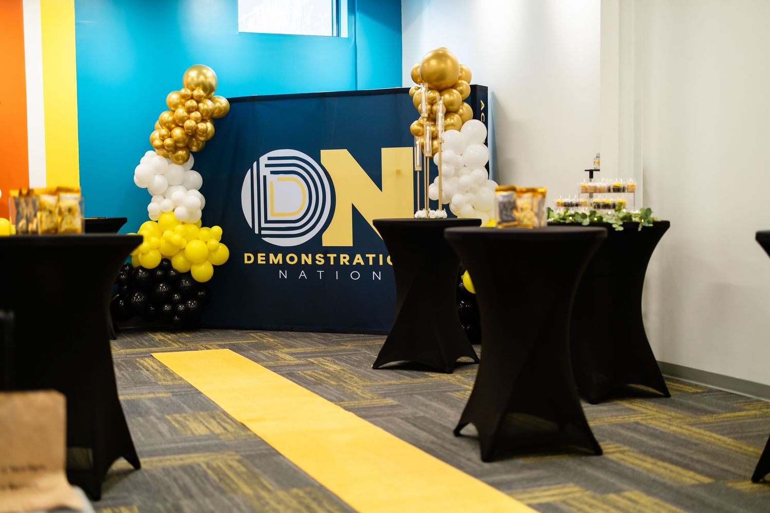 Demonstration Nation branded event setup with gold and white balloon decor, black cocktail tables, and a dessert display.
