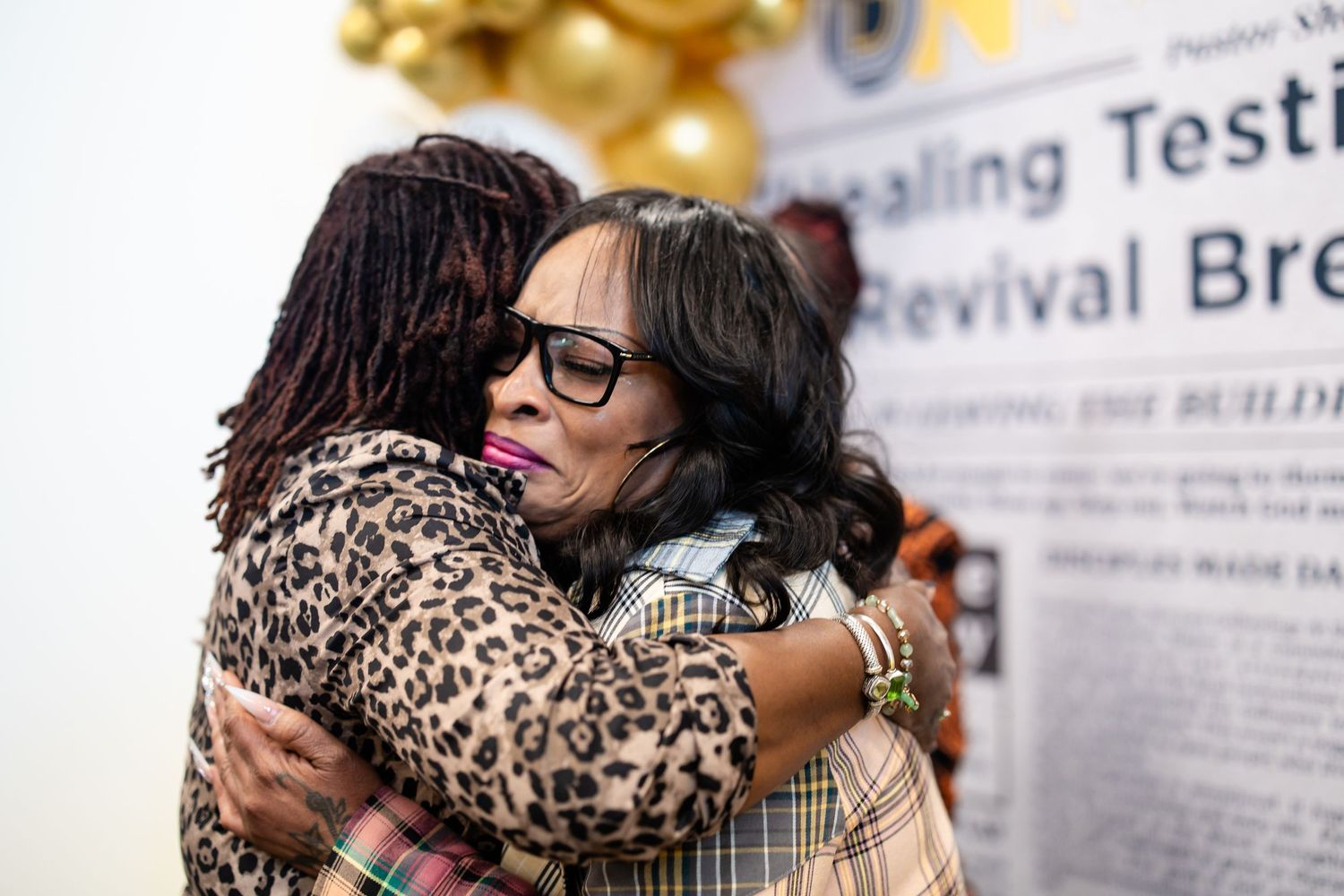 Two women share a warm embrace at a healing testimony revival event with decorative gold balloons in the background.