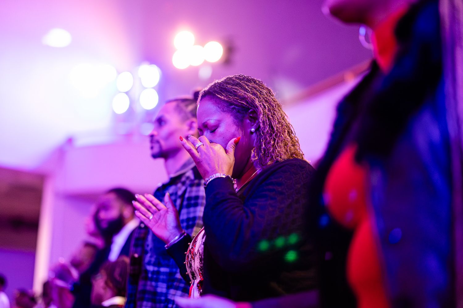 Woman with braids clapping and praying emotionally at a church worship service under purple stage lighting.