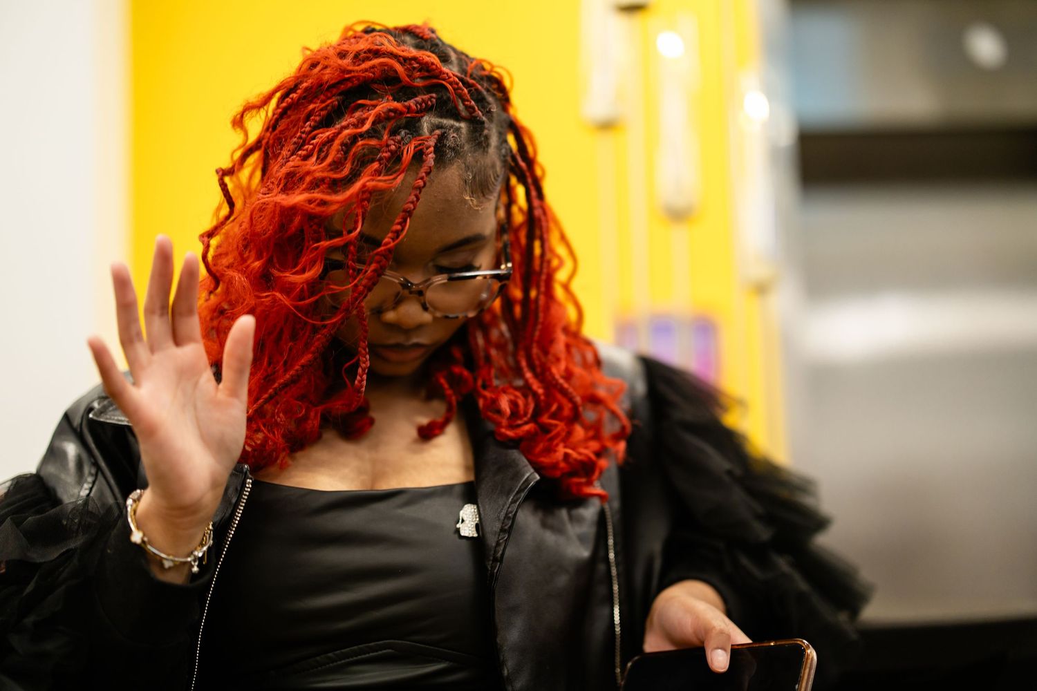 Woman with vibrant red locs wearing a black leather jacket, waving her hand while holding a phone.
