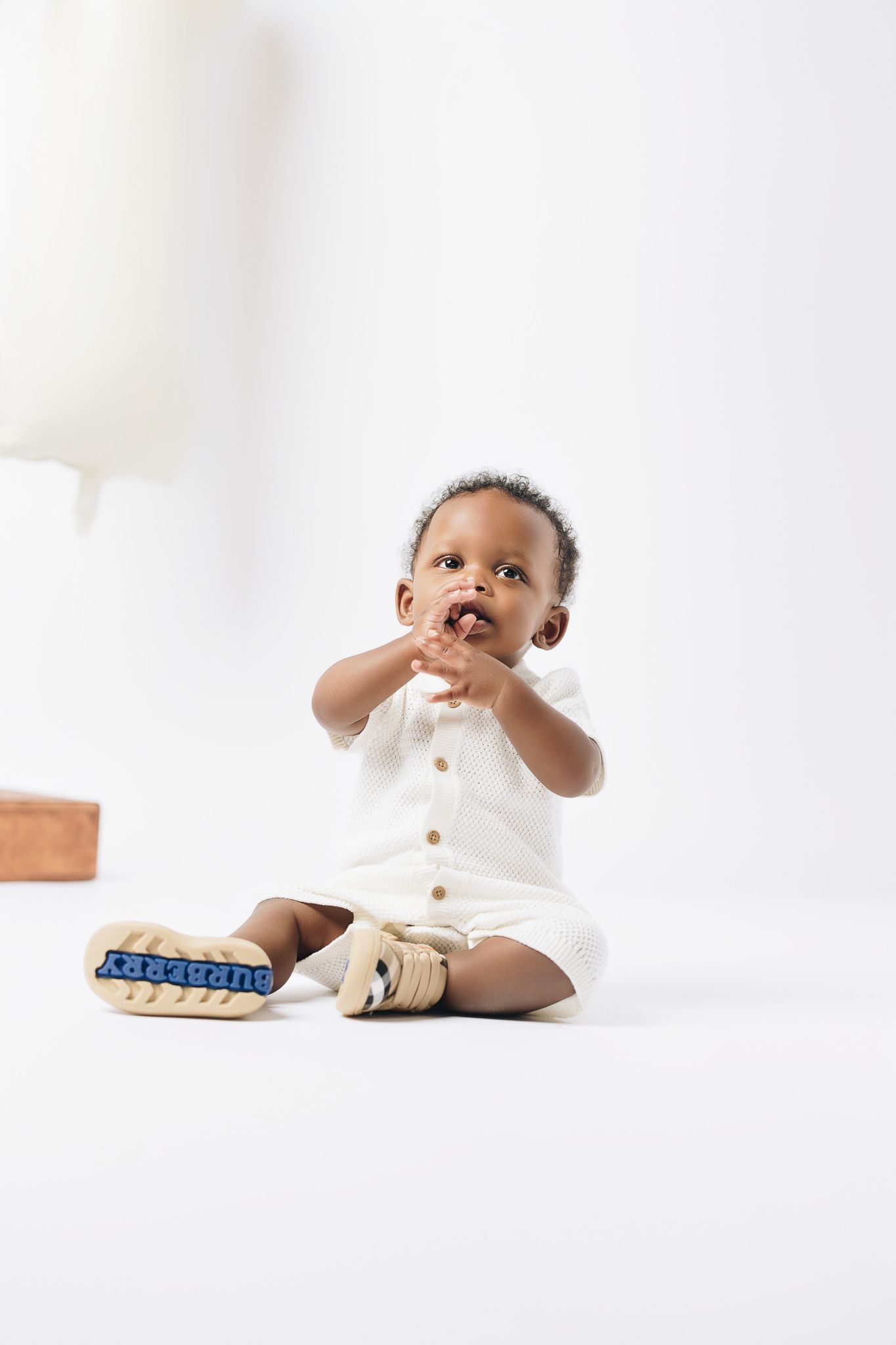 Adorable Black baby in white romper and Burberry shoes sitting on white floor, looking up curiously.