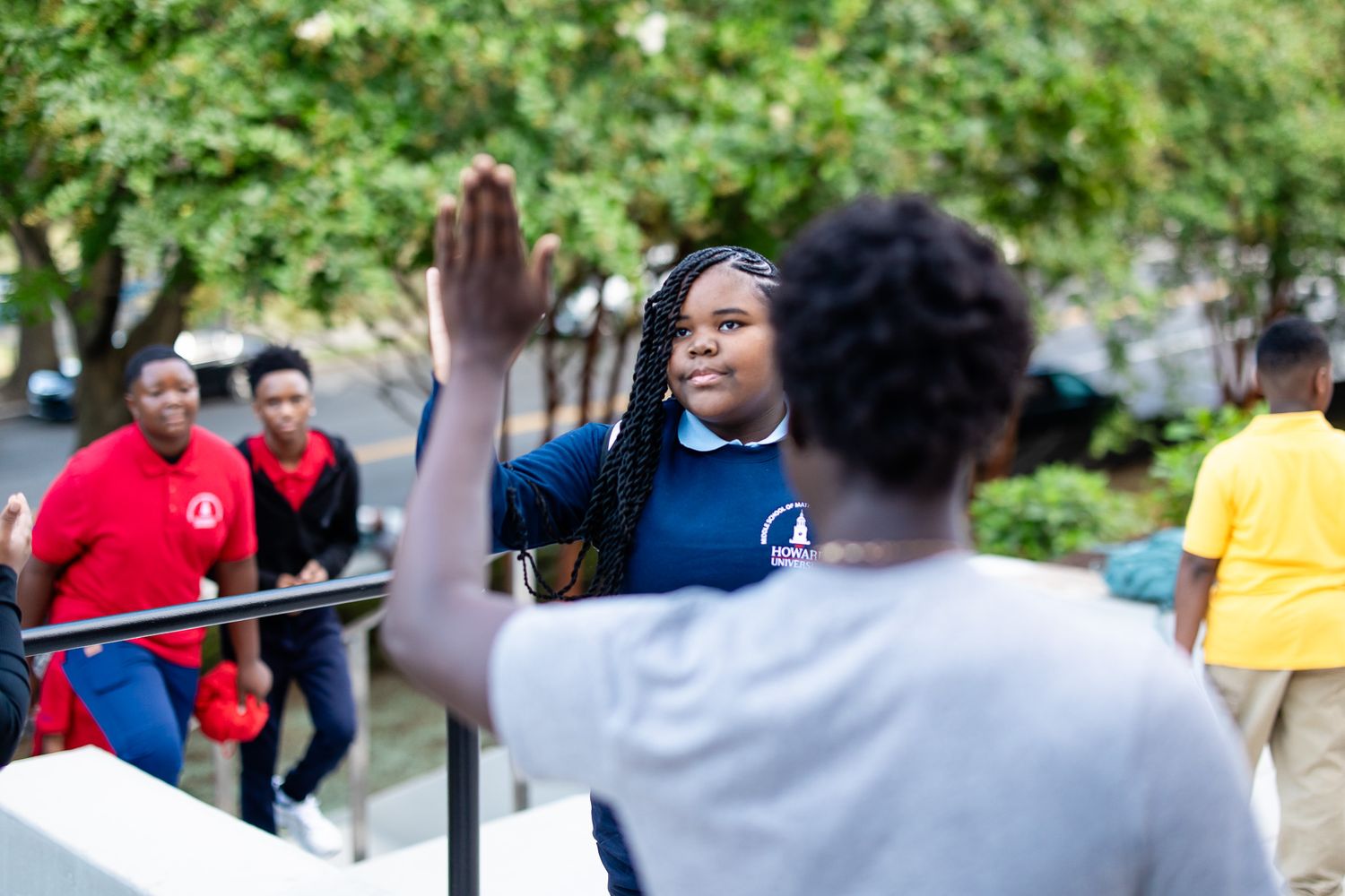 Young Black woman in blue shirt raises hand outdoors, greeting peers near a railing with trees in background.