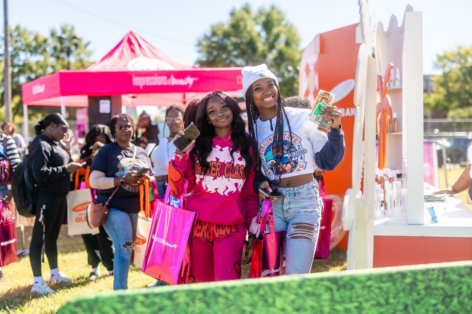 Two women pose together at an outdoor festival near a pink beauty brand tent, holding shopping bags on a sunny day.