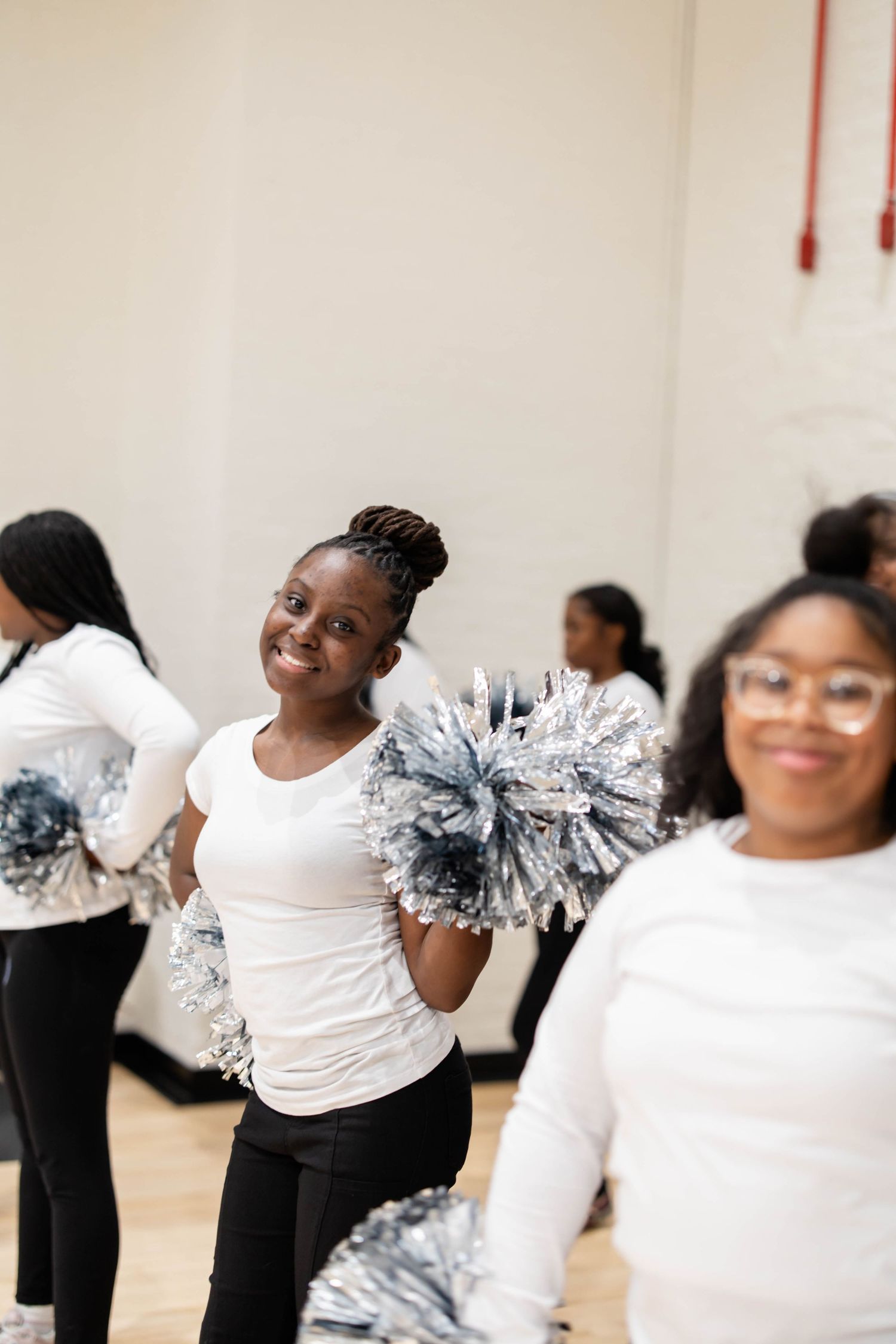 Young cheerleaders in white tops holding silver pom-poms practice routines in a gymnasium, smiling with enthusiasm.