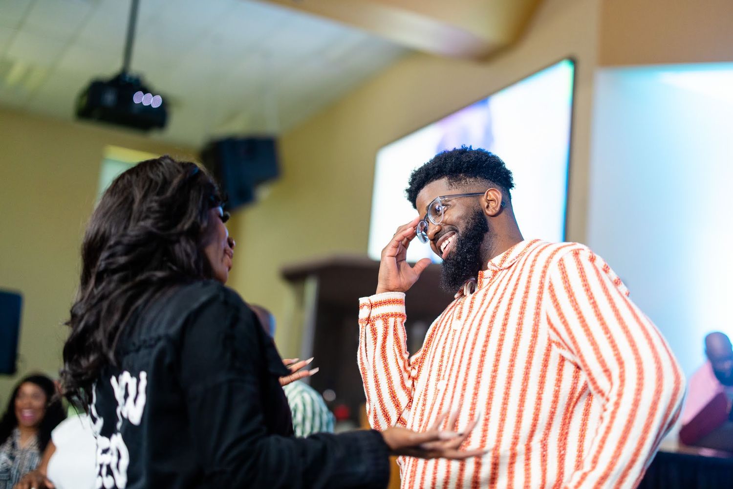 Two people in conversation at an indoor event, man in orange striped shirt adjusting glasses, woman in black jacket.