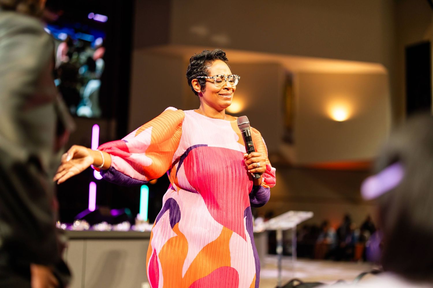 Woman in colorful floral dress holds microphone while speaking on stage at an indoor event.