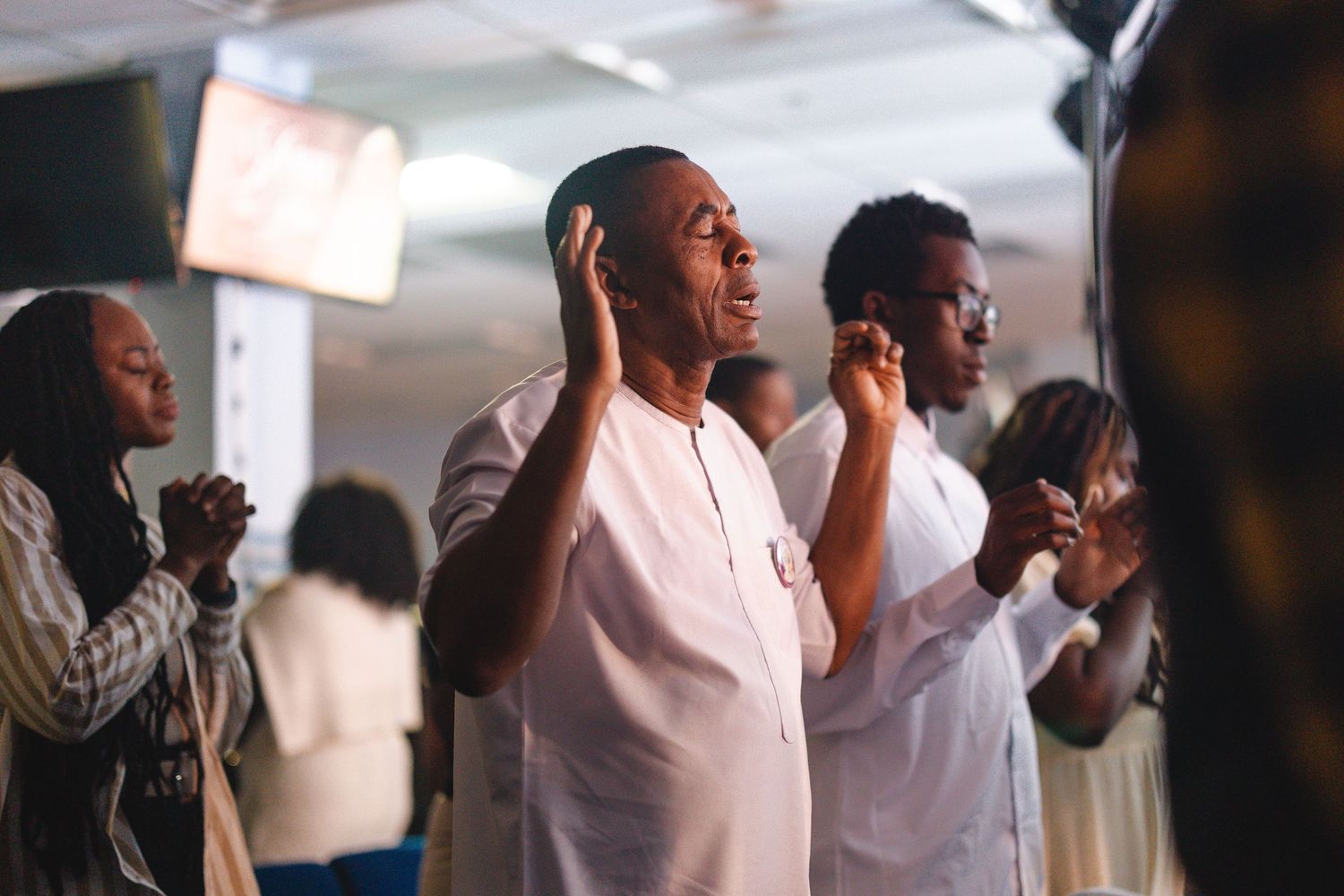 People worshipping in church, with a woman in white raising her hands in prayer during a religious service.