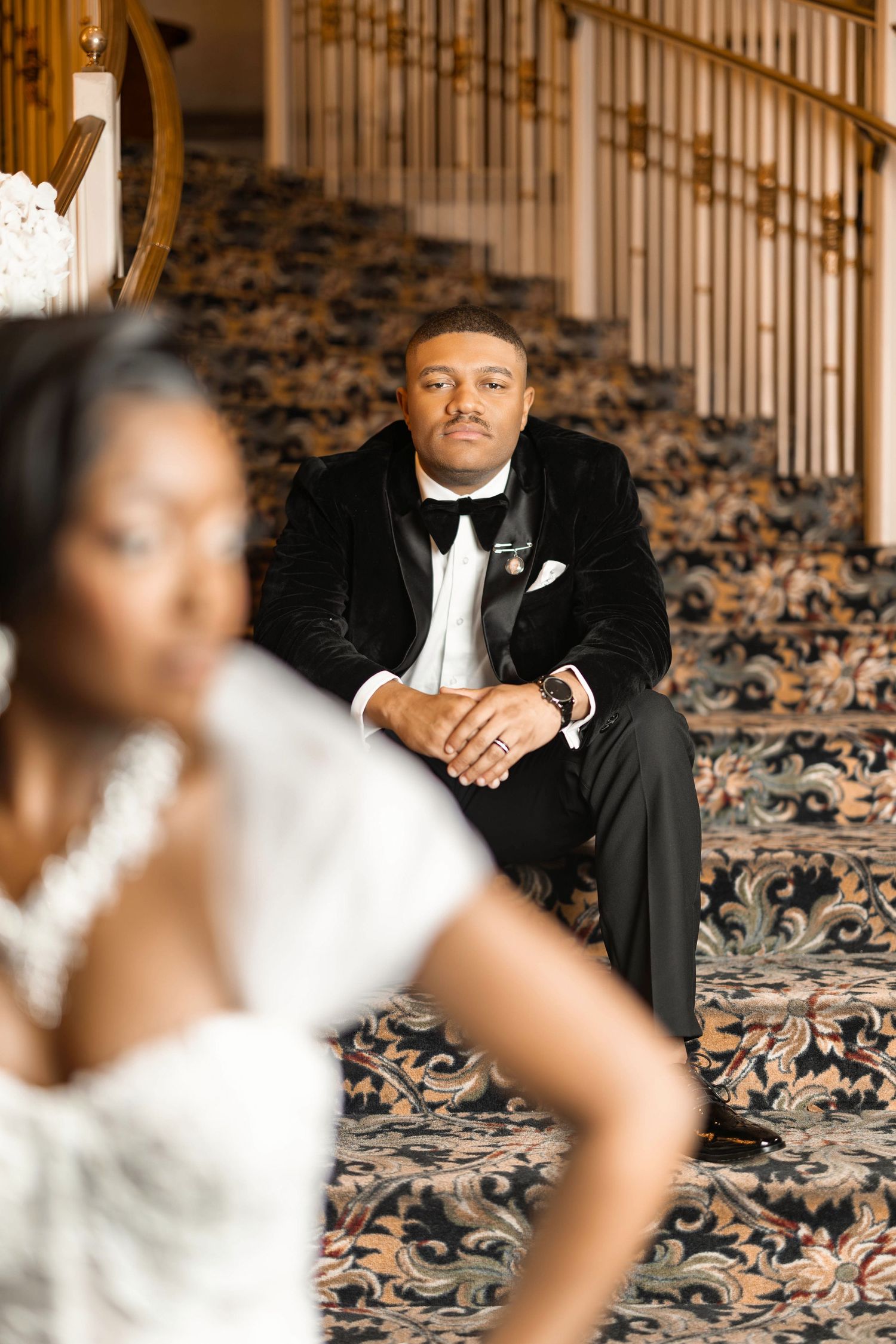 Groom in black velvet tuxedo sits on ornate staircase gazing at bride in white lace dress during wedding portrait.