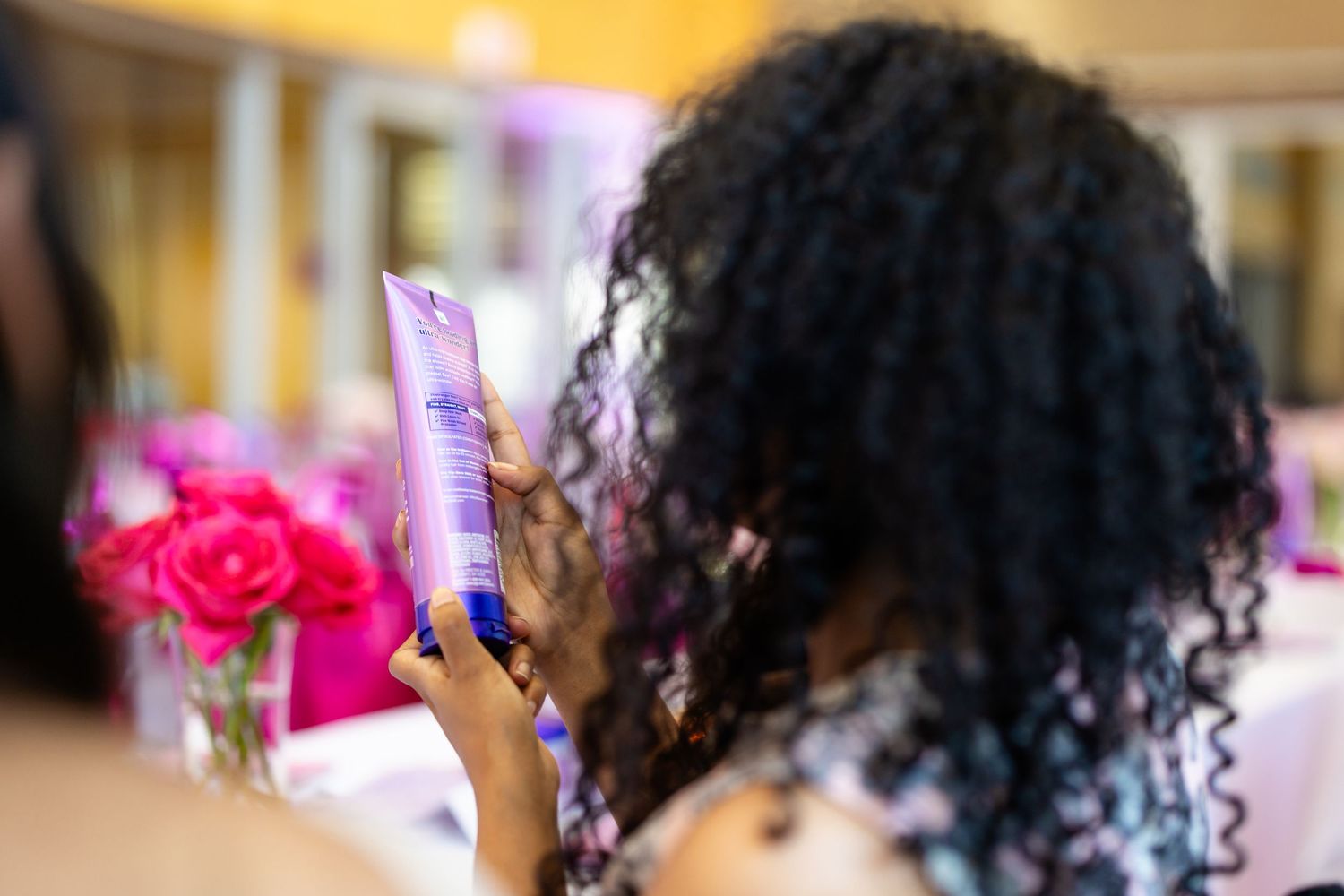 Woman with curly hair reading a purple hair care product label at a pink-themed beauty event.