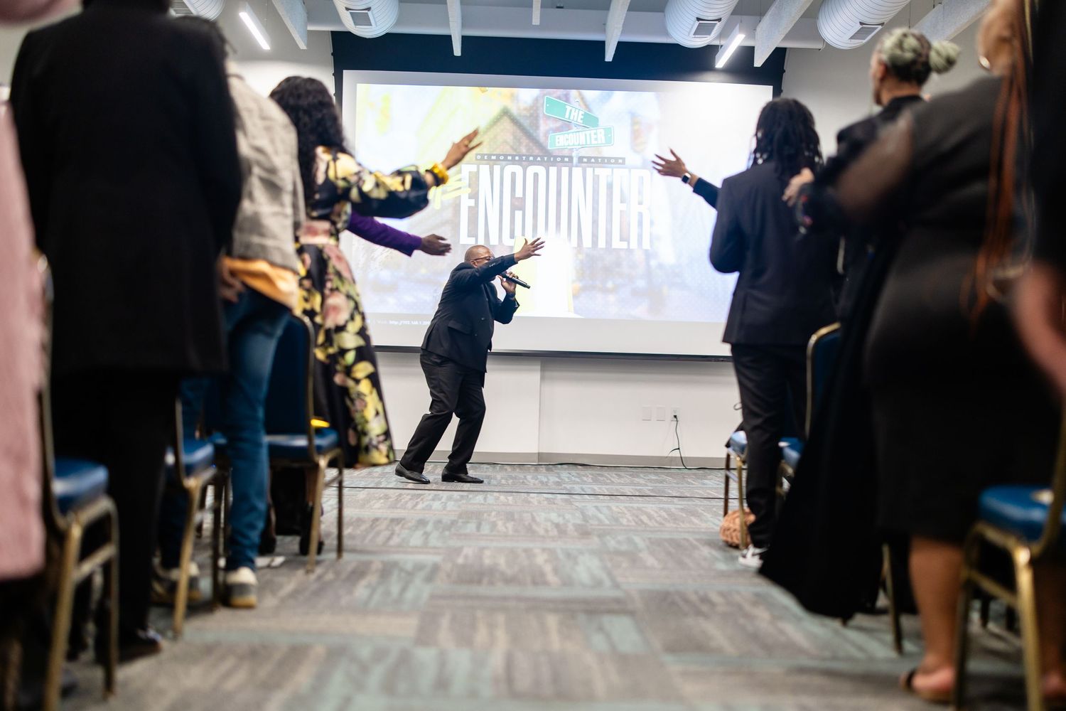 A speaker gestures energetically before a crowd at an Encounters event with a large projection screen backdrop.