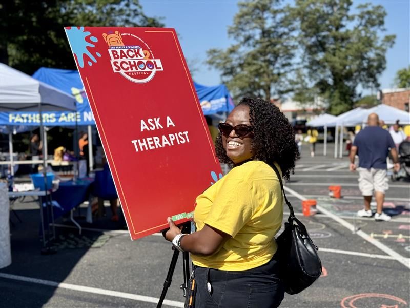 Woman in yellow shirt holds a red Back to School Ask a Therapist sign at an outdoor community event.