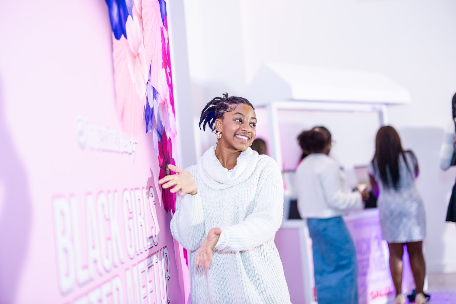 Young woman in white sweater smiling at a Black Girl Magic event display in a bright pink venue.