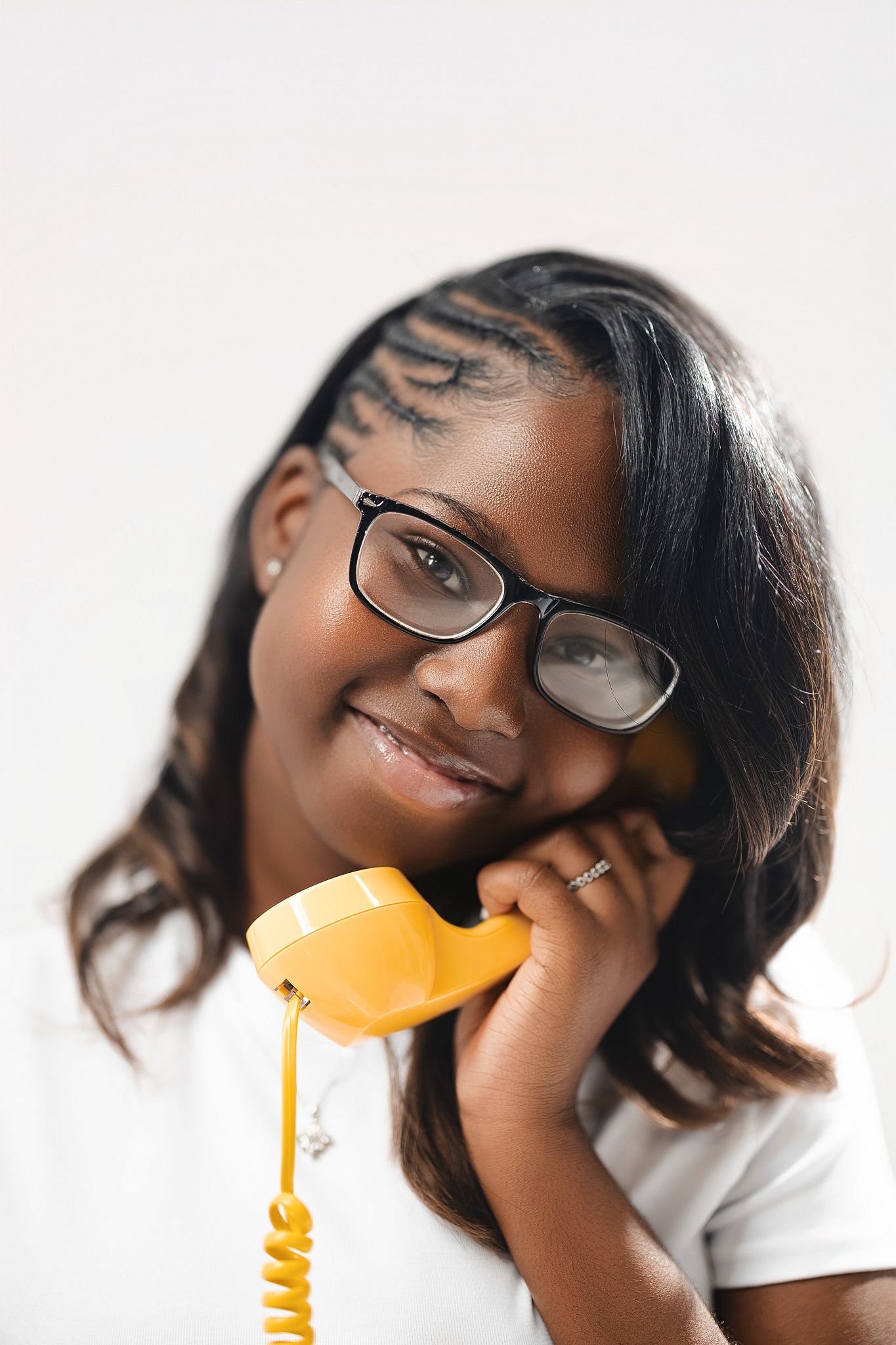 Young girl with braids and glasses smiling while holding a vintage yellow telephone handset against white background.