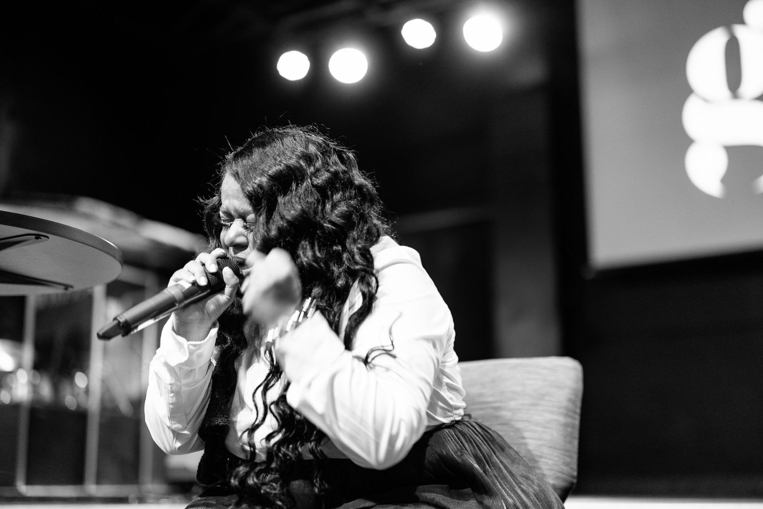 Black and white photo of a woman singing into a microphone on stage, wearing a white blouse and dark vest.