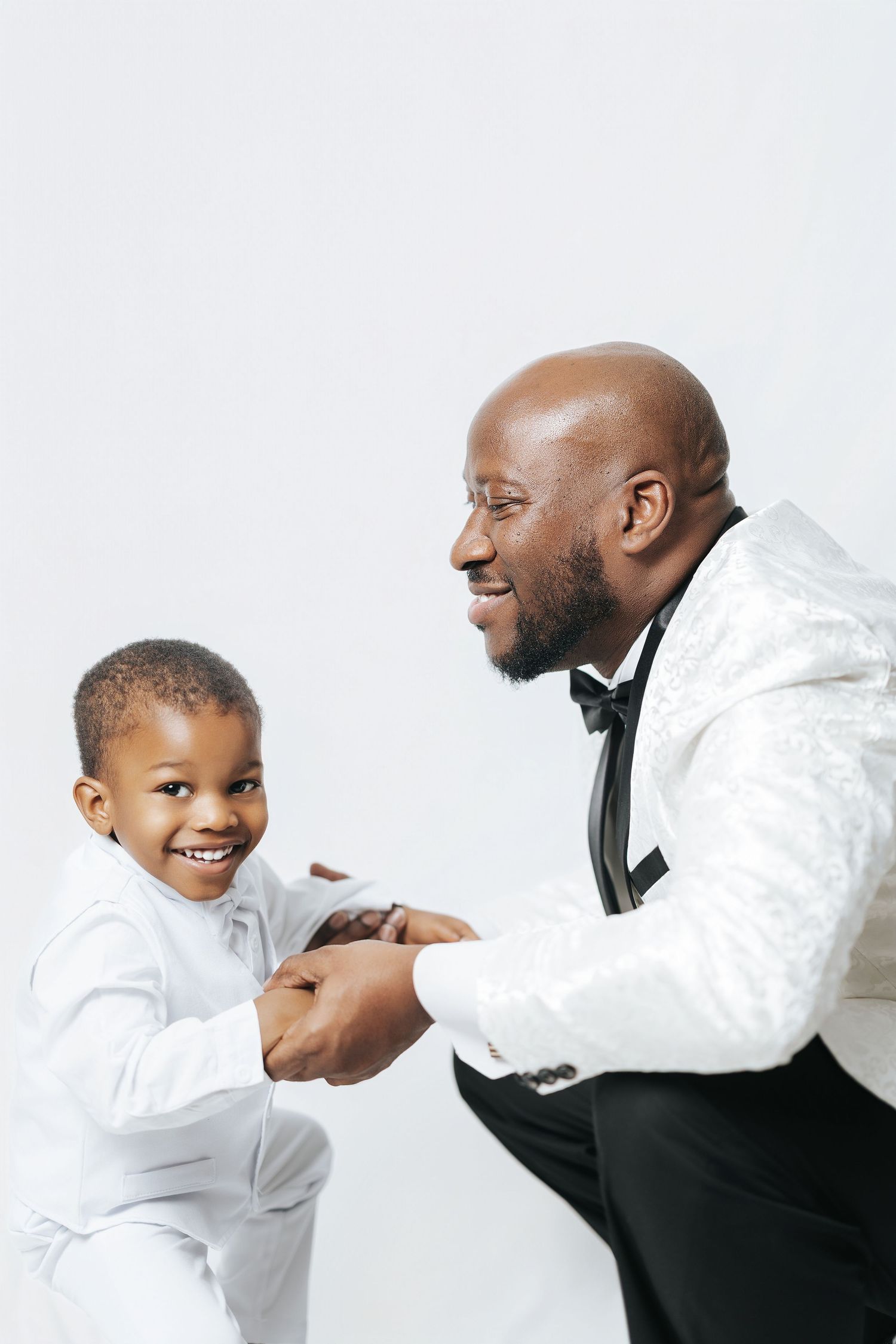 A smiling man in a white tuxedo holds hands with a young boy in a white suit against a light background.
