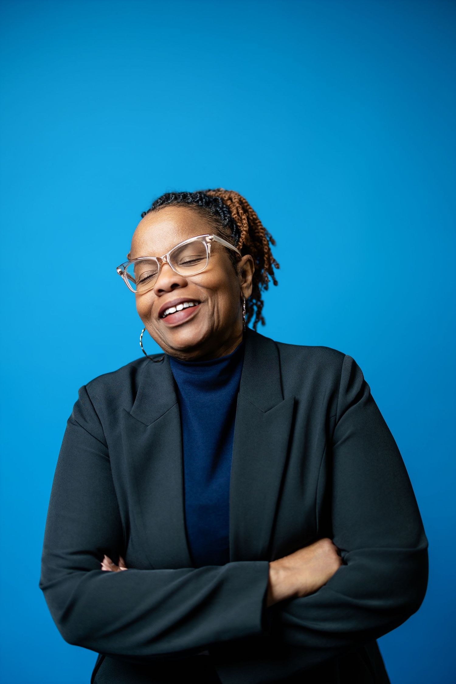 Smiling Black woman with glasses and braids, wearing a dark blazer, arms crossed against a blue background.